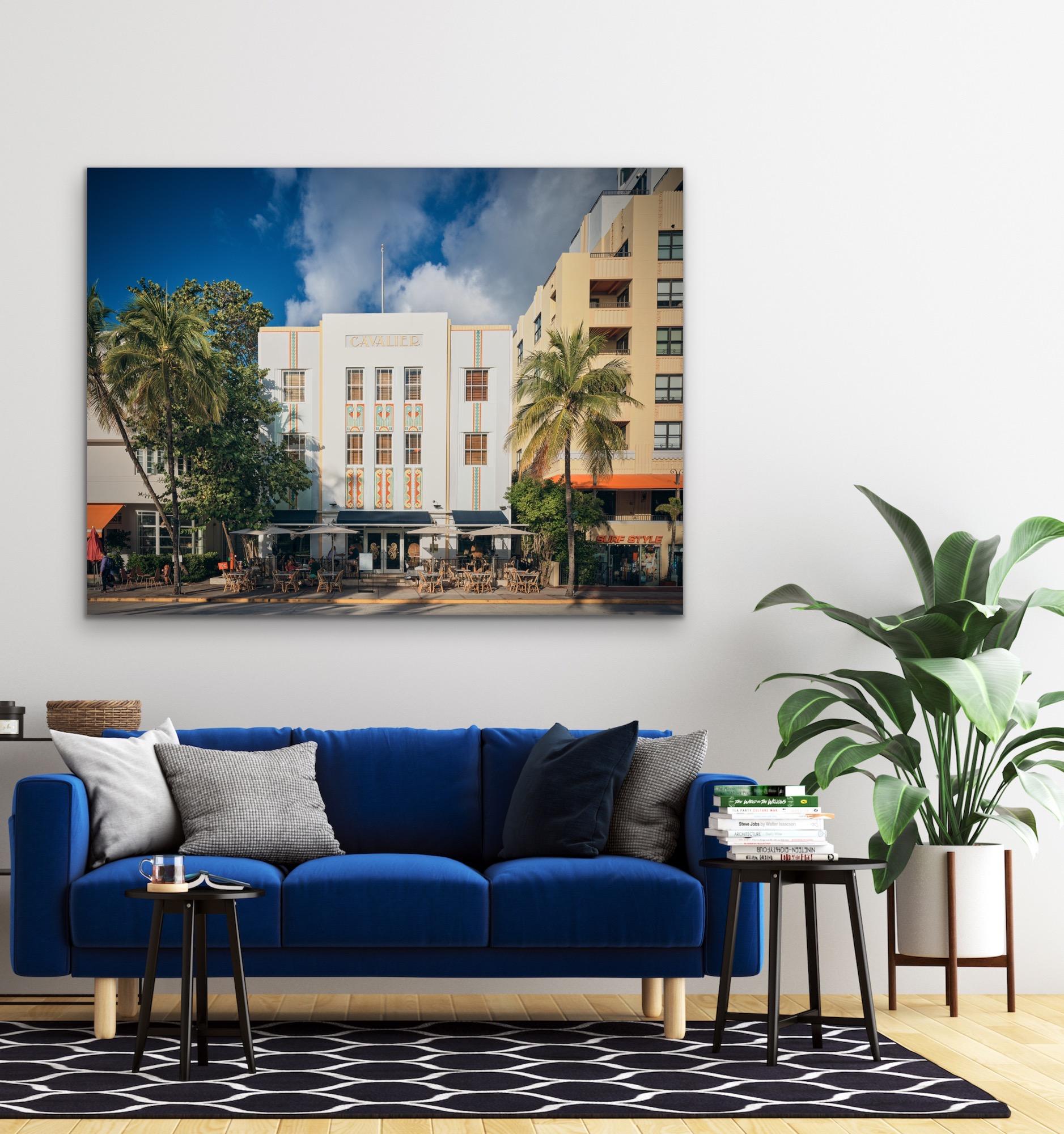 This contemporary urban photograph by Peter Mendelson captures the Deco style Cavalier building in Miami Beach, Florida, with dining tables and umbrellas, and palm trees along the sidewalk. 

This is a metal sublimation print. The frame on an