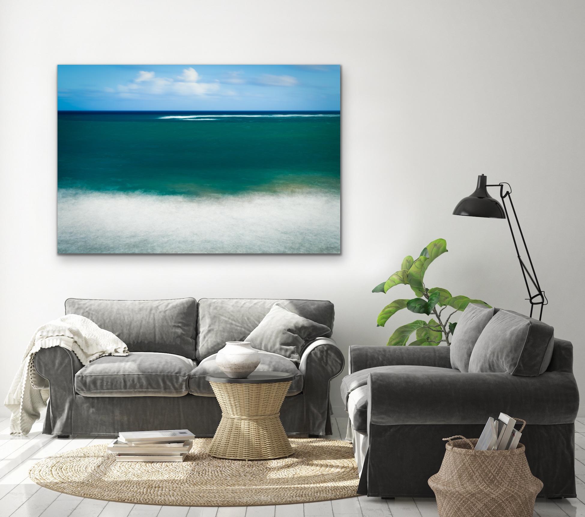 This contemporary coastal photograph by Peter Mendelson is so vibrant and idyllic, it borders on an abstract piece. White, fluffy wisps of clouds float in a bright blue sky above the horizon line of the ocean. Deep blue fades to deep teal, with