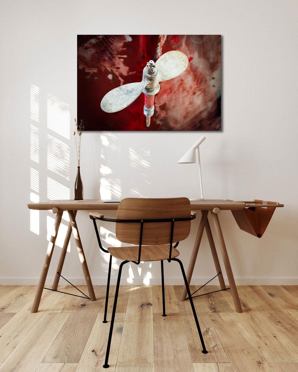 This contemporary coastal photograph by Peter Mendelson features a weathered boat propeller which is in focus and almost appears to float at the center of the composition. It sits in front of a blurred, textured red of the boat, which creates an