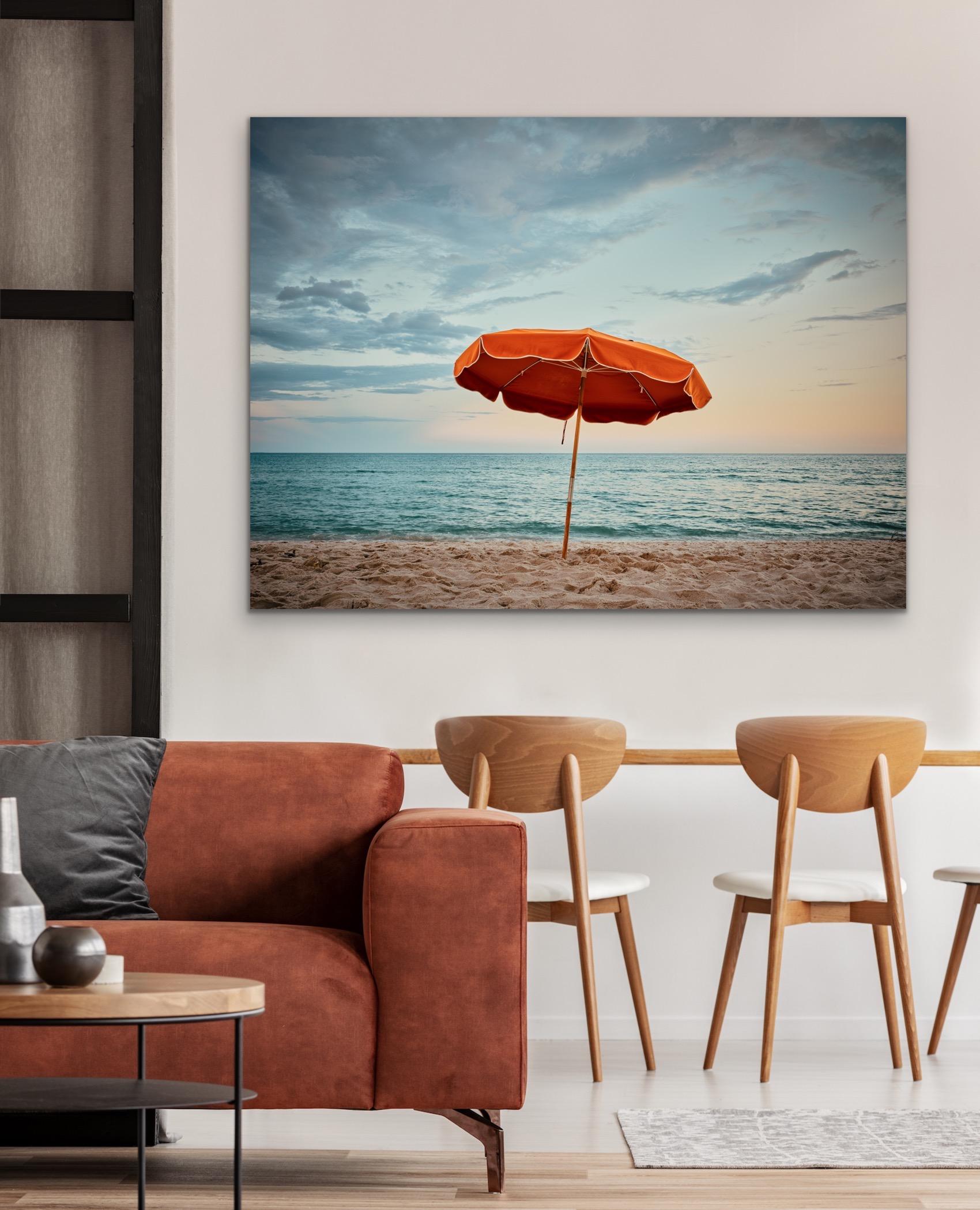 This contemporary coastal photograph by Peter Mendelson captures a vibrant orange beach umbrella planted in sand overlooking the ocean at Miami Beach in Florida. The blue sky has a subtle warm tone along the horizon, tying together the warm tones in