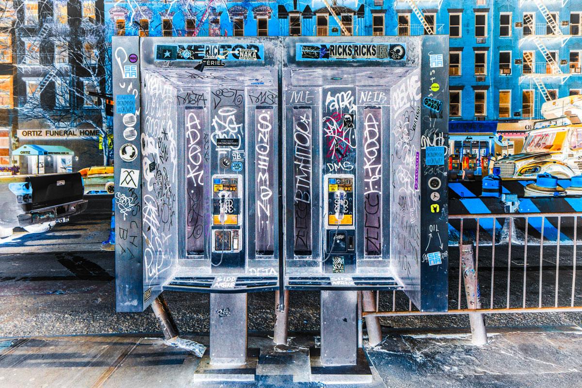 This contemporary urban photograph by Peter Mendelson is a metal sublimation print. It features a cool palette, and captures two phone booths covered in graffiti writing on a city street lined with large buildings. 

This is a metal sublimation