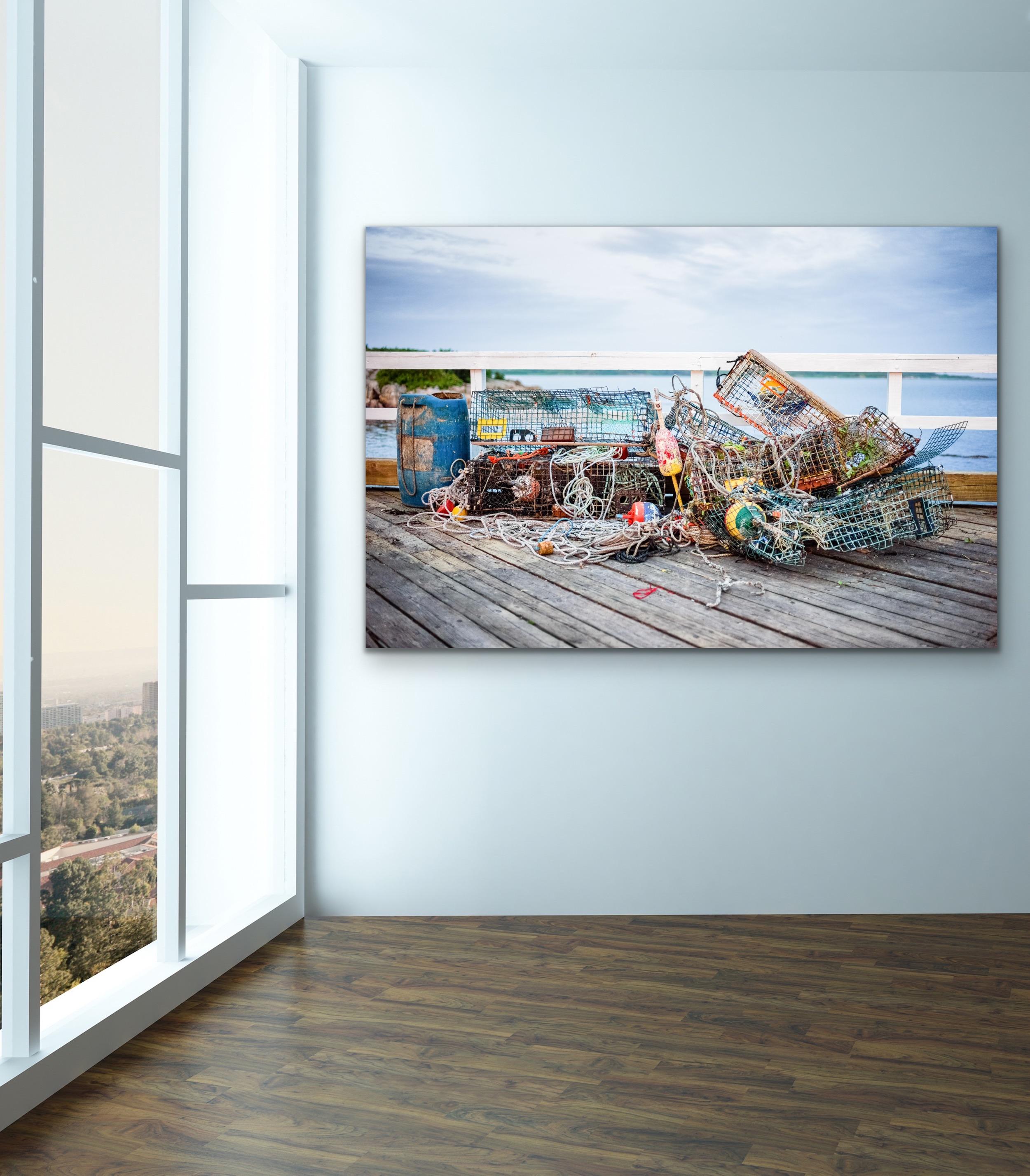 This contemporary coastal photograph by Peter Mendelson pictures colorful lobster traps and nets, stacked in a tangled pile, and sitting on a dock in front of a white railing. Through the railing, the viewer can see a coastal rock formation and the