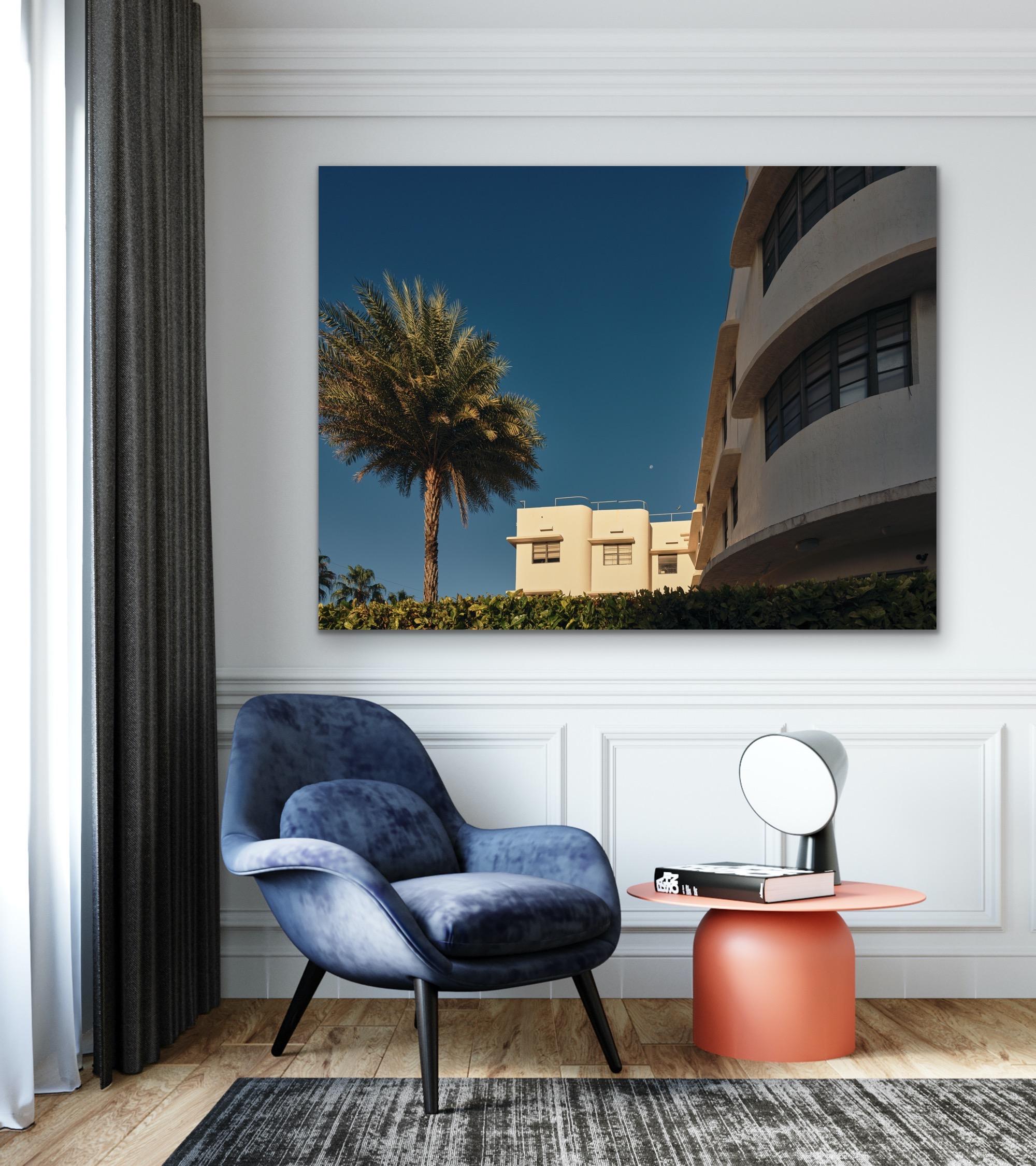 This contemporary coastal photograph by Peter Mendelson captures shadowed buildings next to a palm tree at sunset in Miami Beach, Florida. In the clear blue sky above the white, coastal structures, a small moon is visible. 

This is a metal