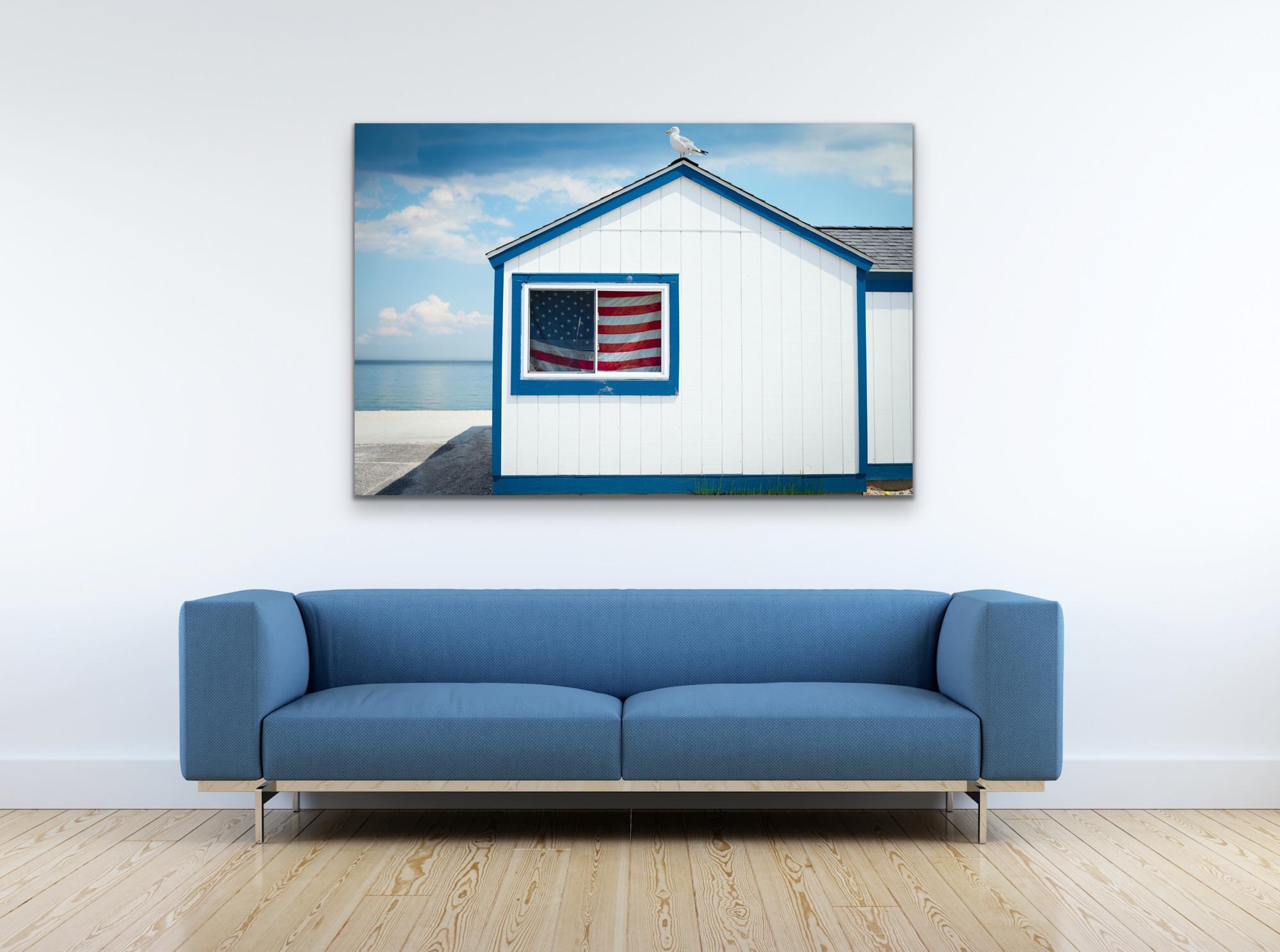 This contemporary coastal photograph by Peter Mendelson pictures a clean, white paneled structure with blue trim. An American flag hangs in the window and a seagull can be seen perched on the peek of the grey roof. The structure sits on concrete