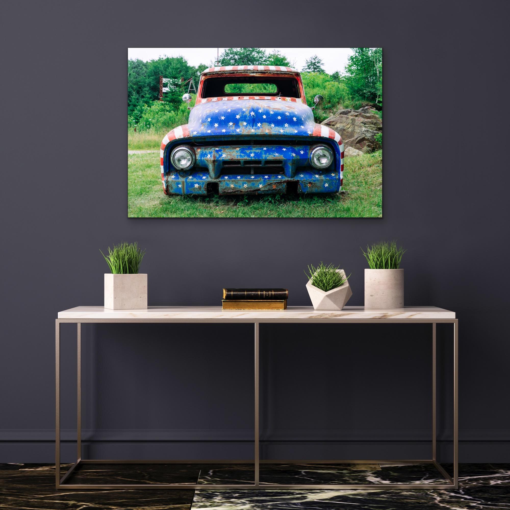 This contemporary photograph depicts an old automobile that has been painted with the pattern of stars and stripes of the American flag. The rusted, window- and tire-less car seems to have been left in a forested, rural area. The photograph was