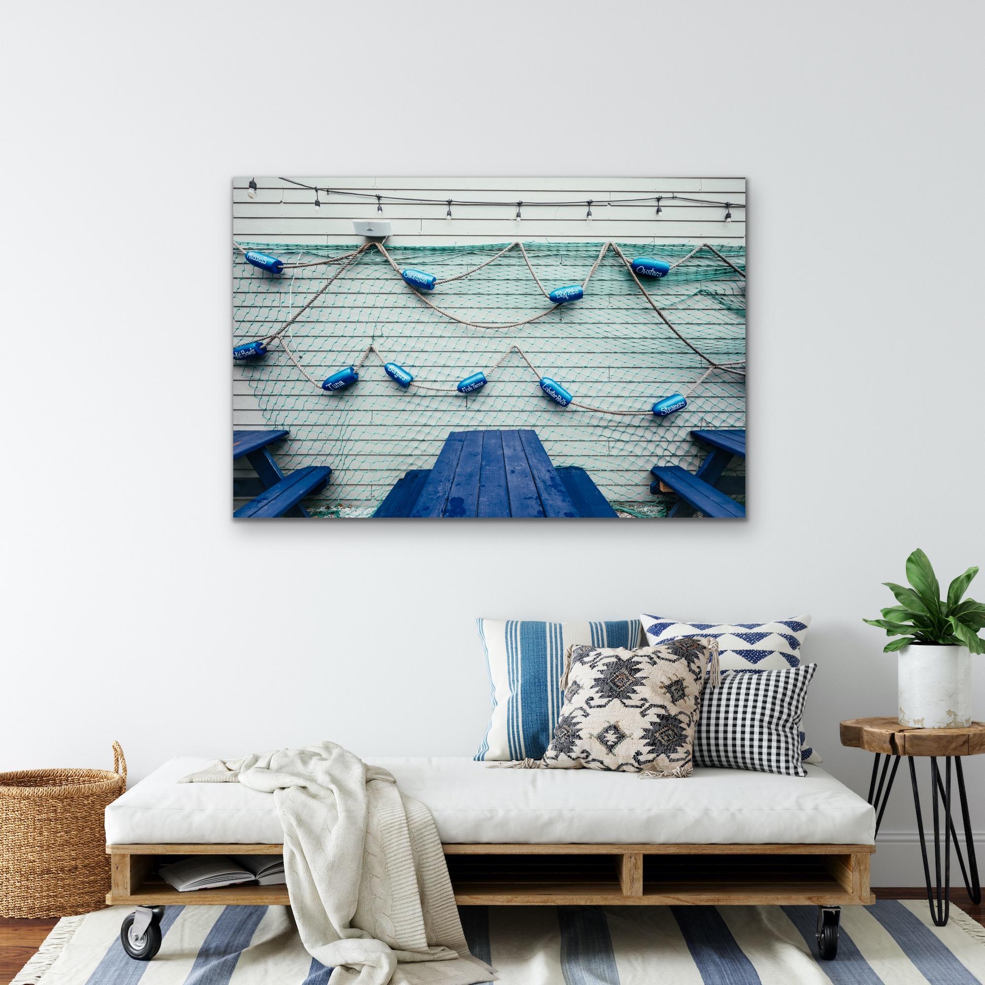 This contemporary coastal photograph has a cool blue palette. It captures a cropped view of blue picnic tables in front of the side of a building that has blue fishing net, buoys, and twinkle lights decorating the vinyl panelling. 

This is a metal