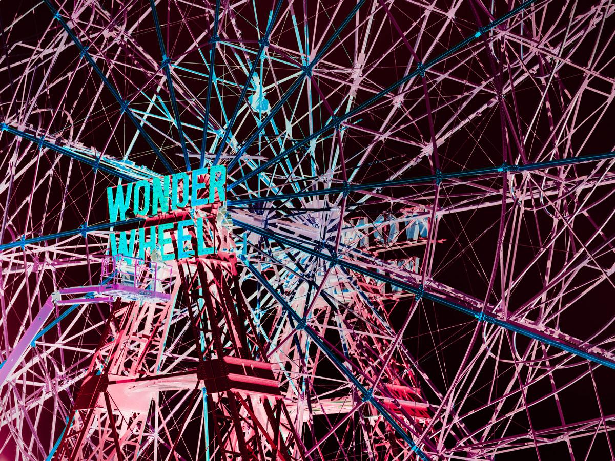 This contemporary photograph by Peter Mendelson captures a large ferris wheel from an angle, lit up with pink and blue lights and a sign that says 