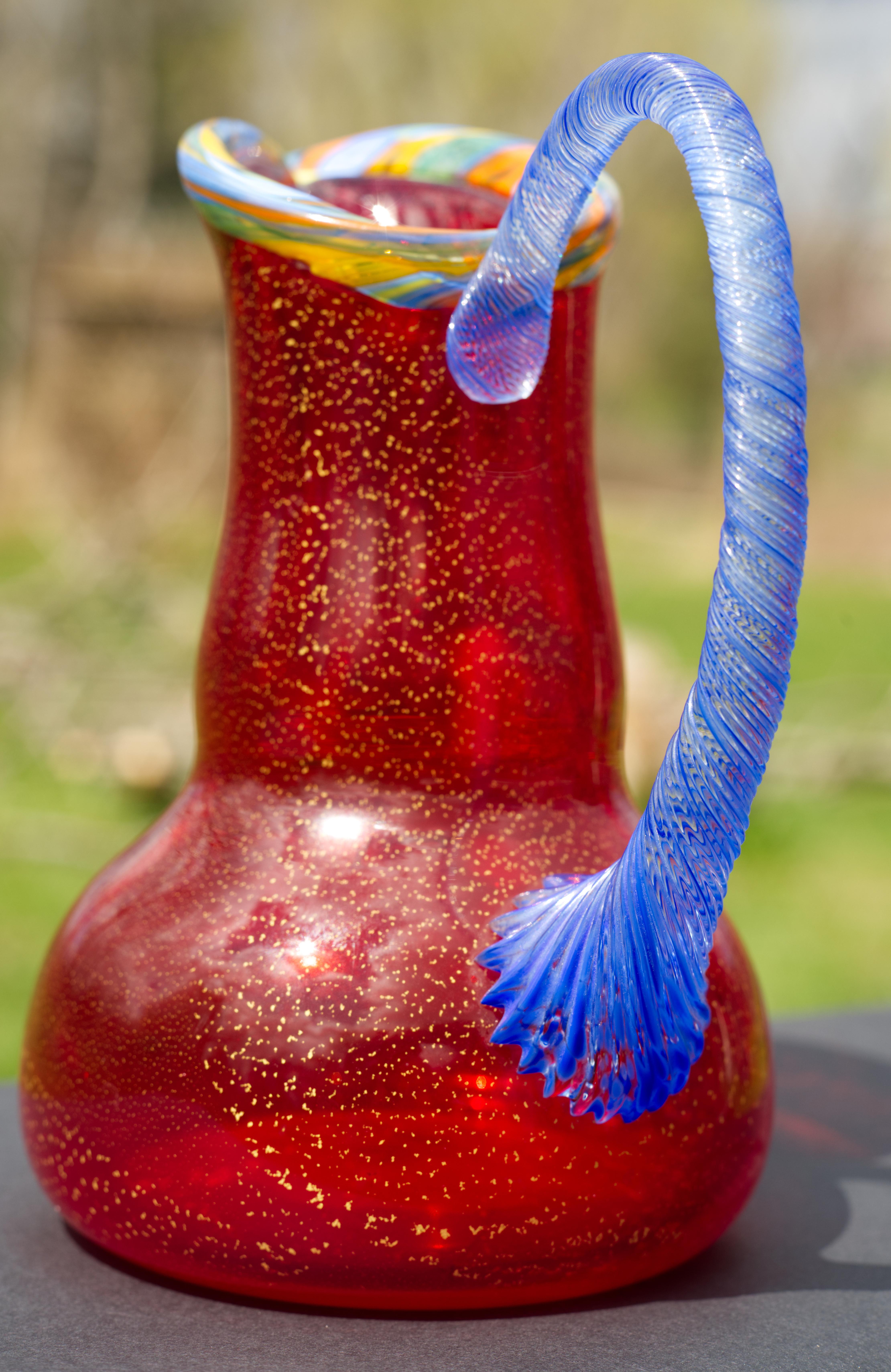 
The pitcher or vase was hand blown by Peter Secrest in Naples, NY. It has ruby red body with gold flakes inclusions throughout; it is decorated with applied textured blue and clear glass handle and striated multicolored collar. The bright, cheerful