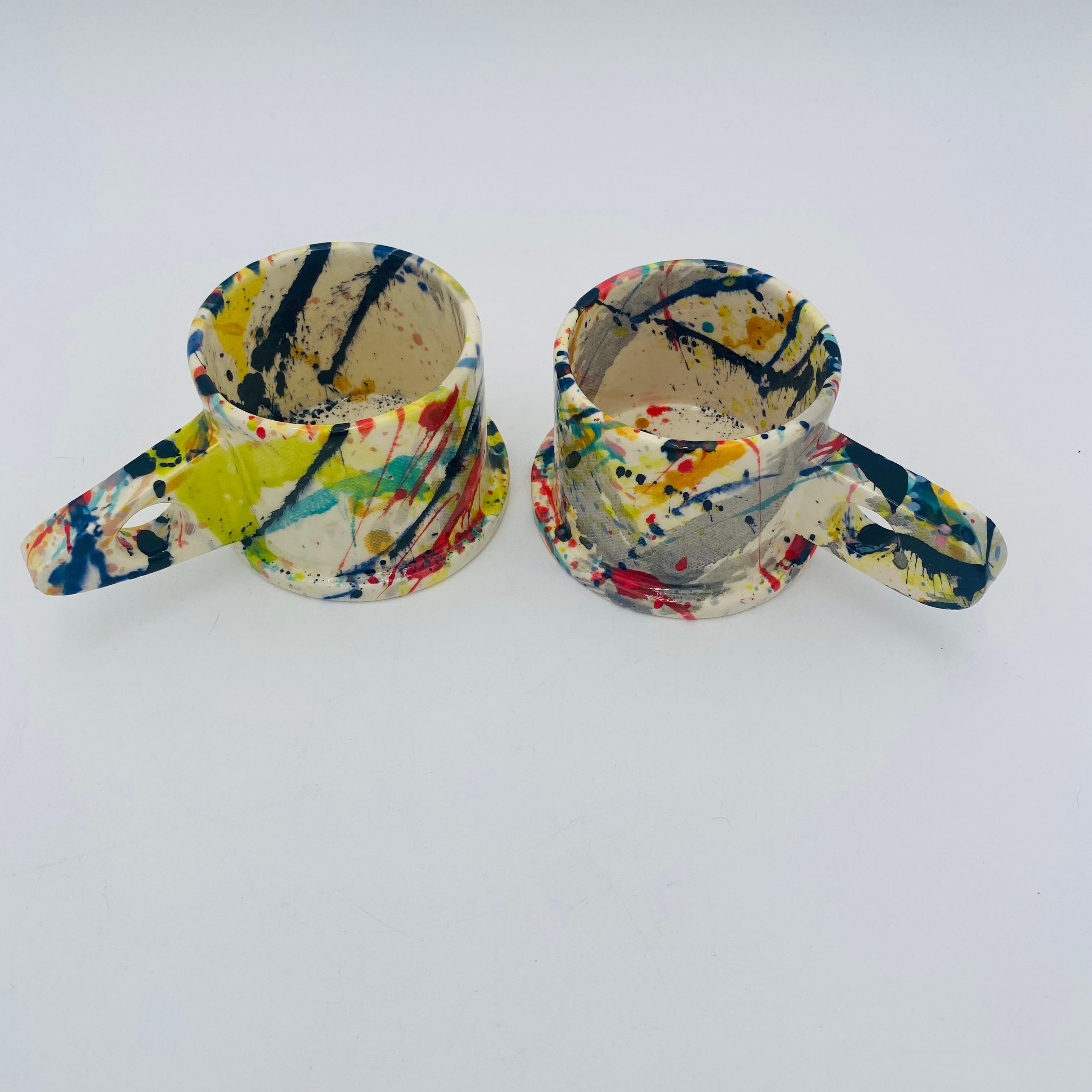 This is a set of two handmade ceramic mugs with multicolored paint splatter by veteran Memphis artist Peter Shire. They are hand-painted, so no two are the same. At the base, each mug reads EXP 2018 in stamped relief text. 

Peter Shire is an