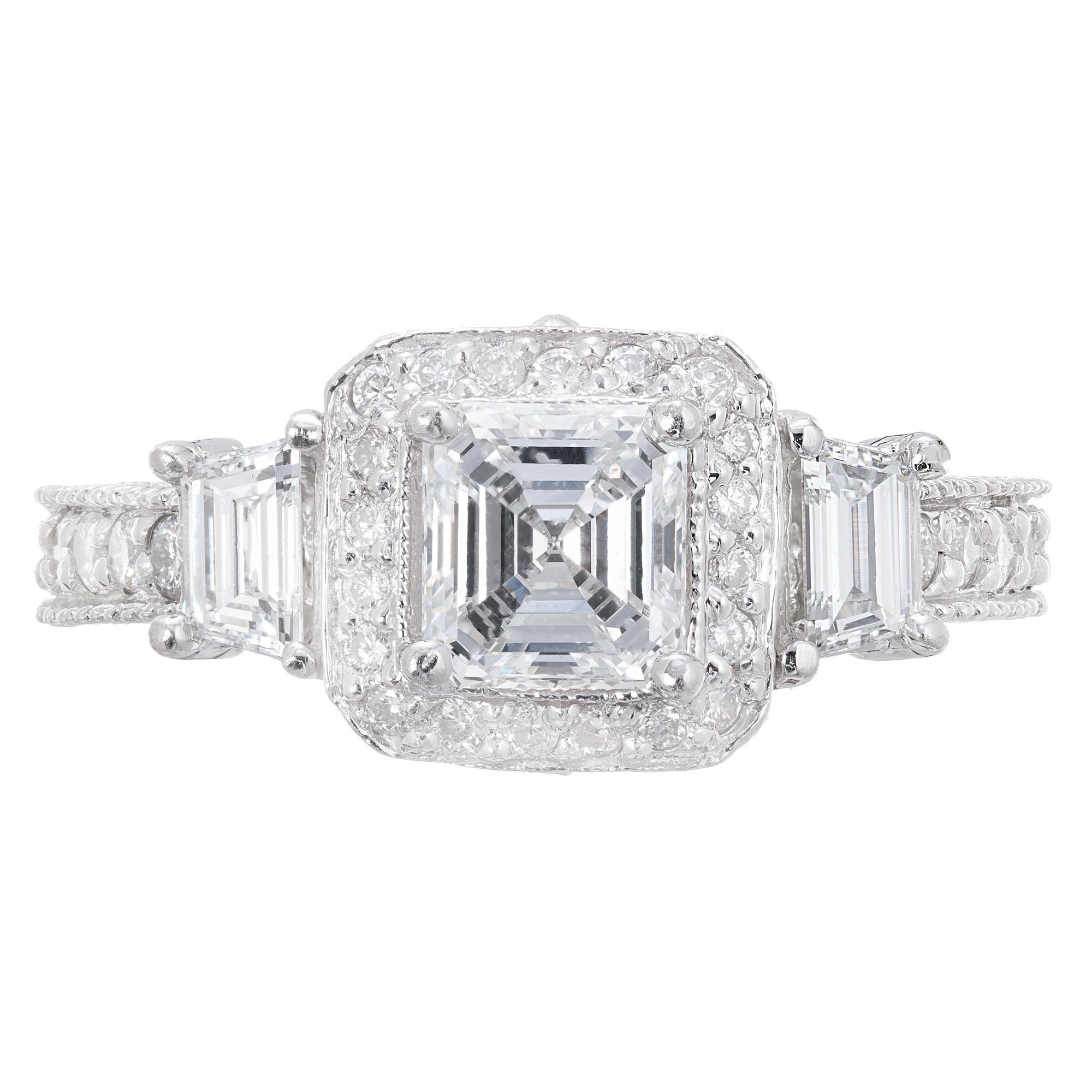 Diamond three-stone engagement ring. GIA certified 1.01 Asscher cut center stone with a halo of round cut diamonds. Accented with 2 trapezoid side diamonds and round cut diamonds along the hand engraved shank. Designed by Peter Suchy Workshop.