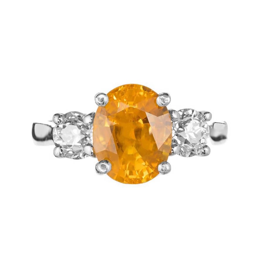 This luxurious ring features a breathtaking 3.29 carat orange sapphire, exuding a warm, vibrant glow reminiscent of a sunset. This captivating gemstone is gracefully embraced by two old European cut shimmering diamonds, one set on each side of the