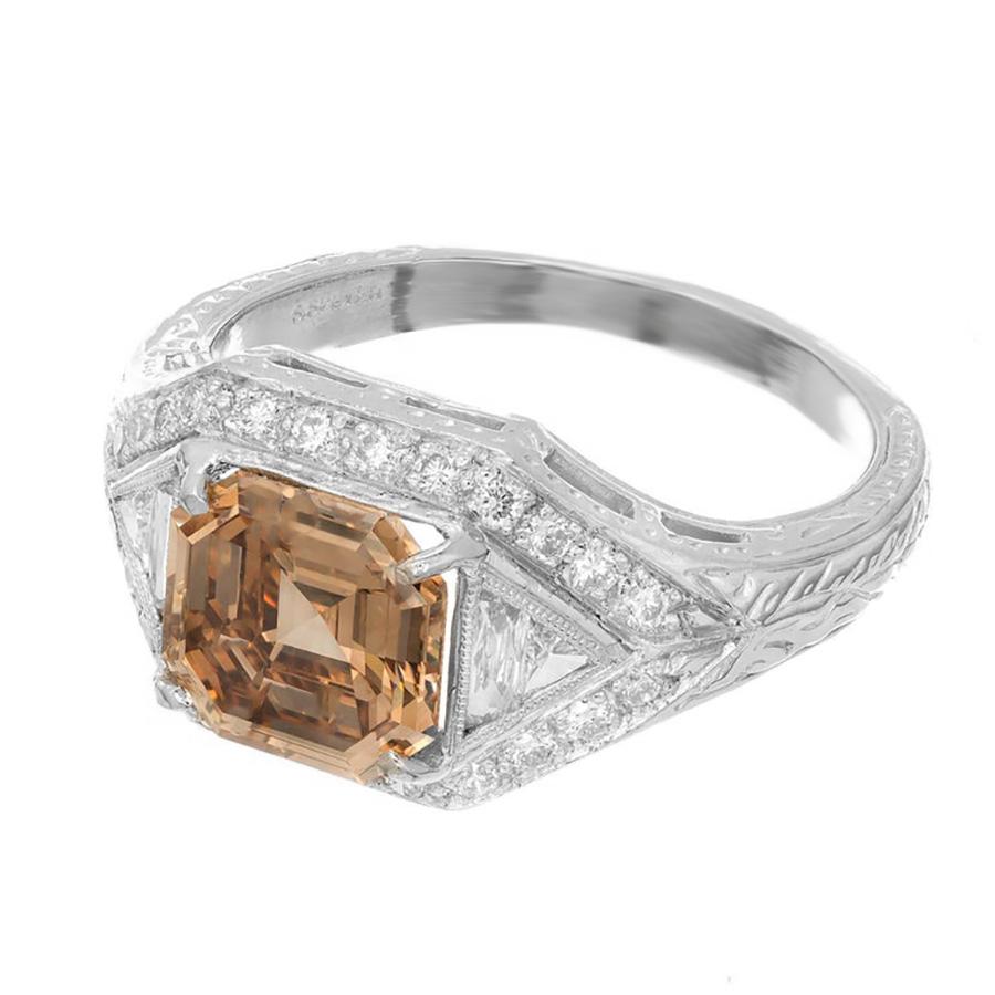 Natural 2.00ct Asscher cut brown and yellow diamond halo engagement ring. The diamond is accented by 2 trapezoid and triangle cut diamonds with a halo of round full cut diamonds. The platinum setting has hand engraving along both sides of the shank.