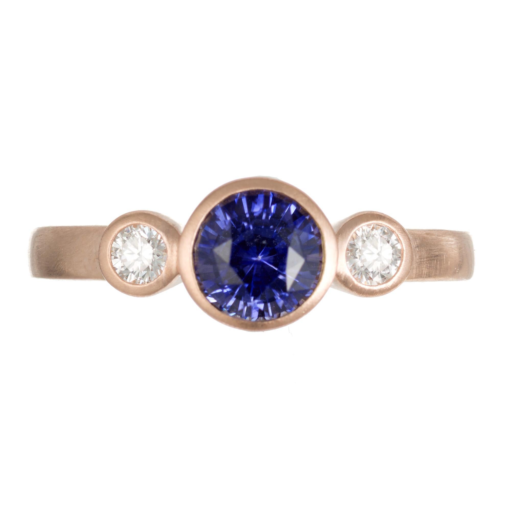 Sri Lanka, Ceylon sapphire and diamond engagement ring. This ring begins with a .96ct round center sapphire which is mounted in a 14k rose gold bezel setting. The rich blue and violet sapphire is accented buy one round brilliant cut diamond on each