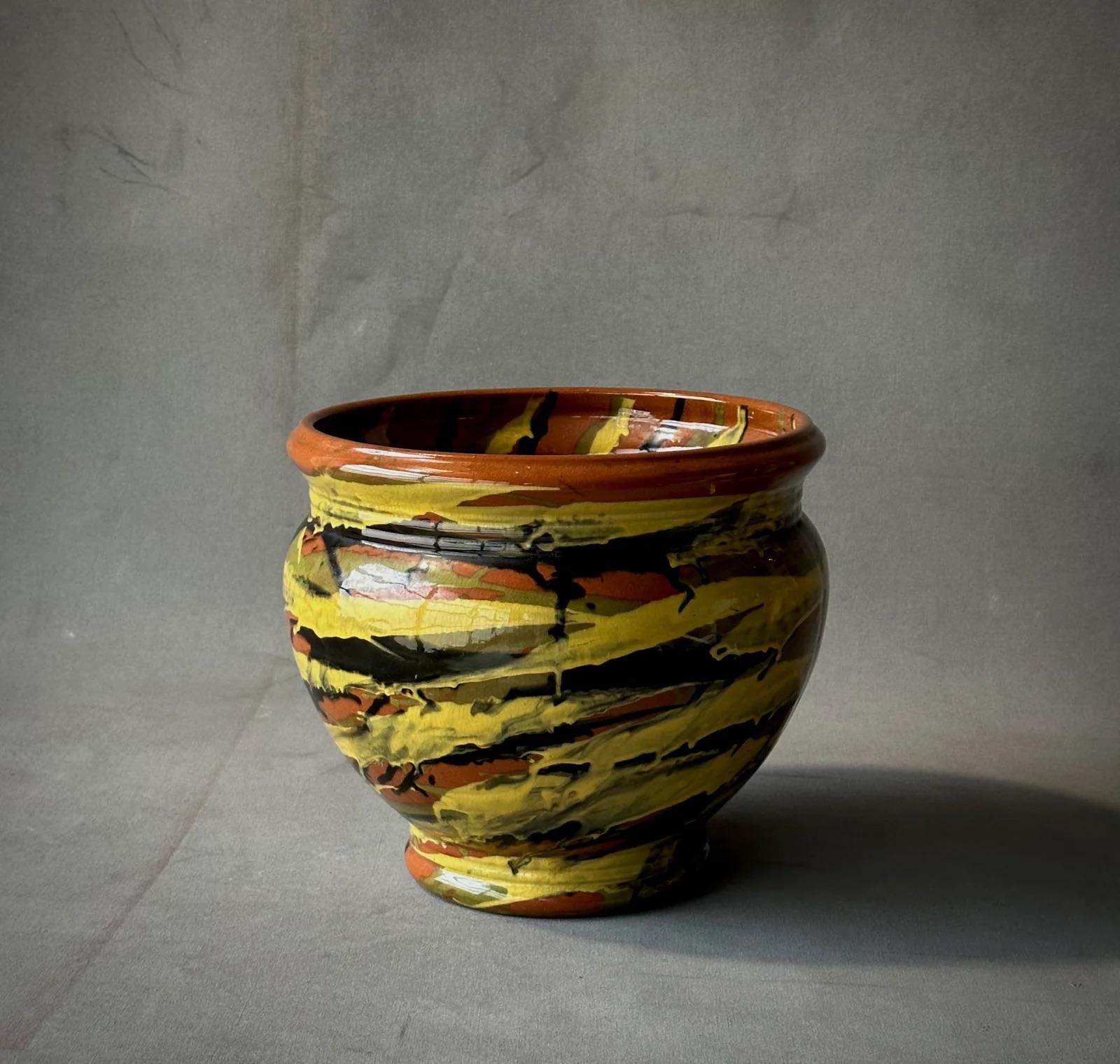 This multi-colored ceramic pot by Peters & Reed exudes a vivid palette of mustard, ink, moss, and terracotta, crafted with thick, energetic daubs of glaze that accentuate its dynamic, swirling form. Originating from Zanesville, Ohio, this piece is a