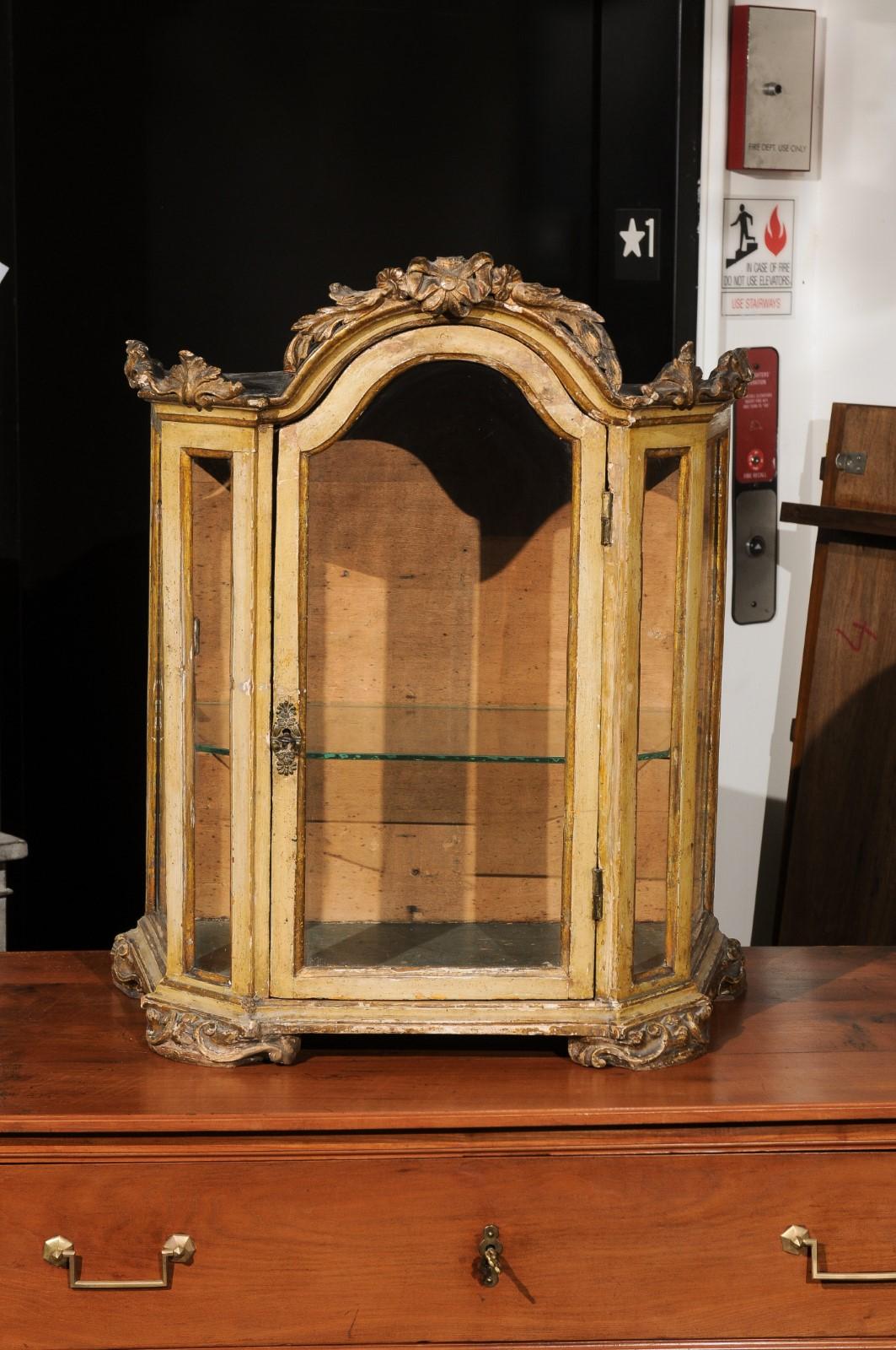 A petite Venetian 18th century period Rococo painted wood vitrine with carved crest and glass door. Created in Italy during the Rococo period, this Italian table top vitrine features an exquisite ribbon-tied floral carved crest, elegantly resting on