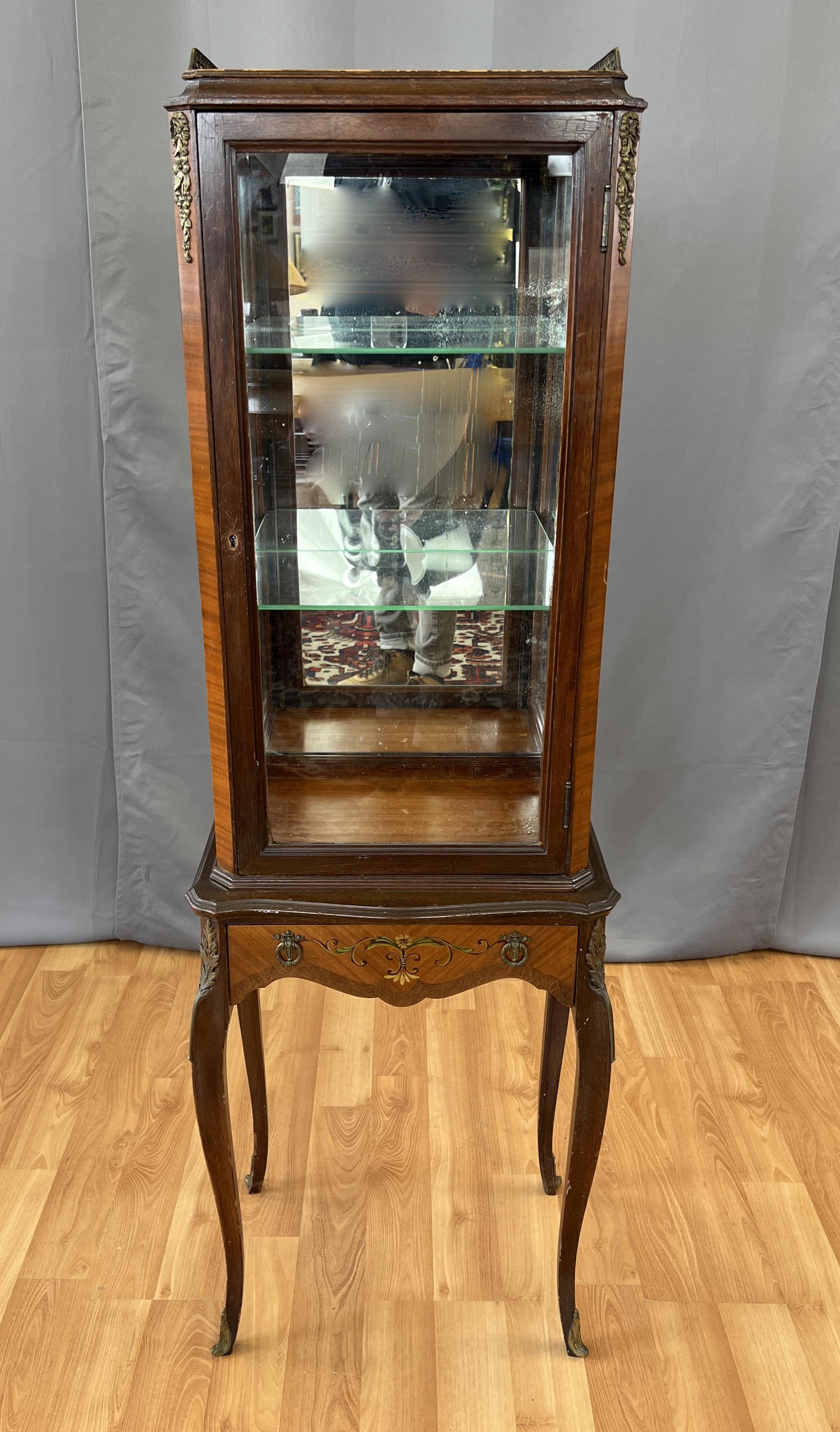 A wonderful circa 1920s Demi Edwardian Vitrine in the style of Louis XVI. Three sided glass unit on four long cabriole style legs finished with caps. Expertly hand painted florals on small drawer with two decorative pulls. Large glass wooden framed