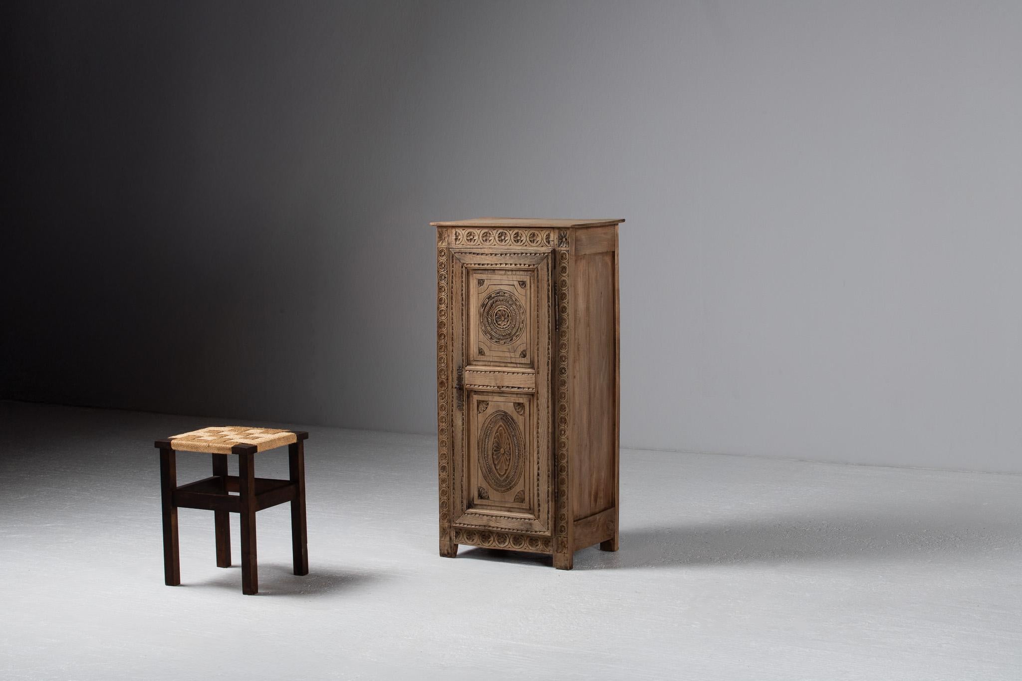 Solemn and ceremonial, this petite armoire stands like a devotional column—its oak façade incised with rhythmic motifs that echo both liturgical ornament and rural craftsmanship. The carved reliefs unfold in measured cadence: floral rosettes, radial