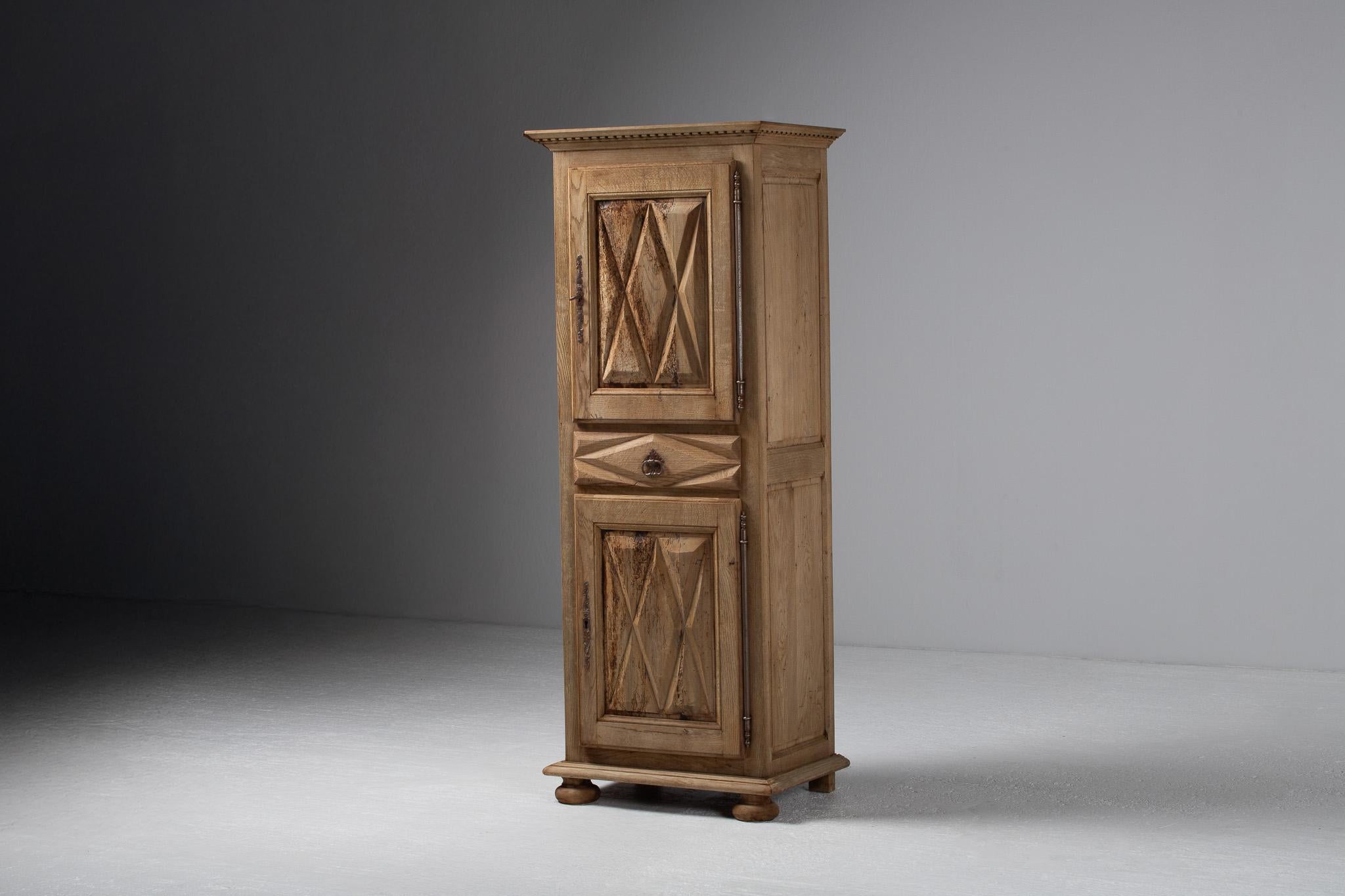 Esbelto y arquitectónico, este pequeño armario se alza con el porte de una columna: estrecho en su postura, pero rico en detalles. Sus puertas gemelas están talladas en relieve con puntas de diamante afiladas, cada faceta atrapa la sombra como lino