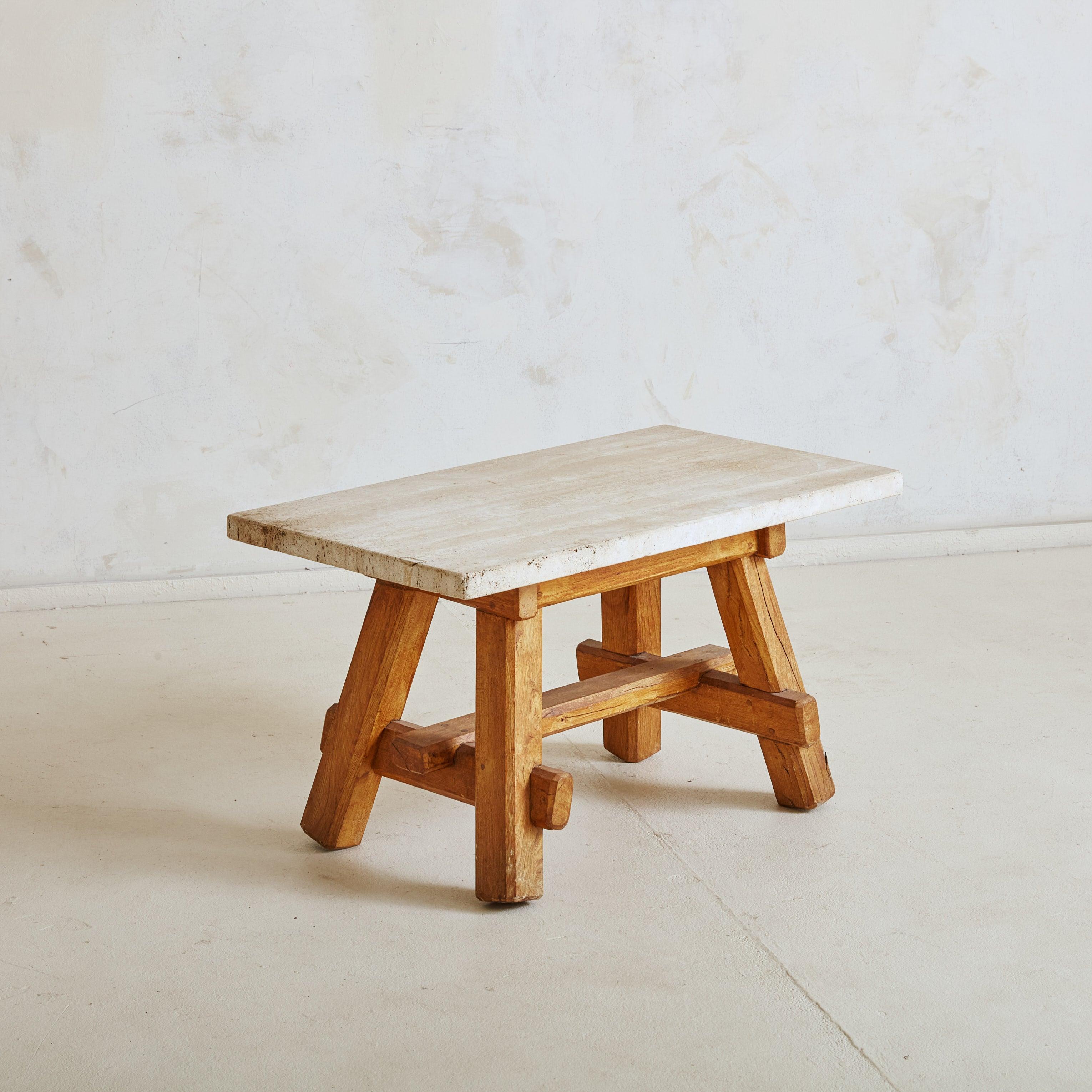 A petite 1950s French hand-carved wooden coffee table featuring sculptural legs and a bottom stretcher with mortise and tenon joints. A smooth, rectangular travertine top in hues of beige, milky white, and taupe rests upon the Brutalist-style base.