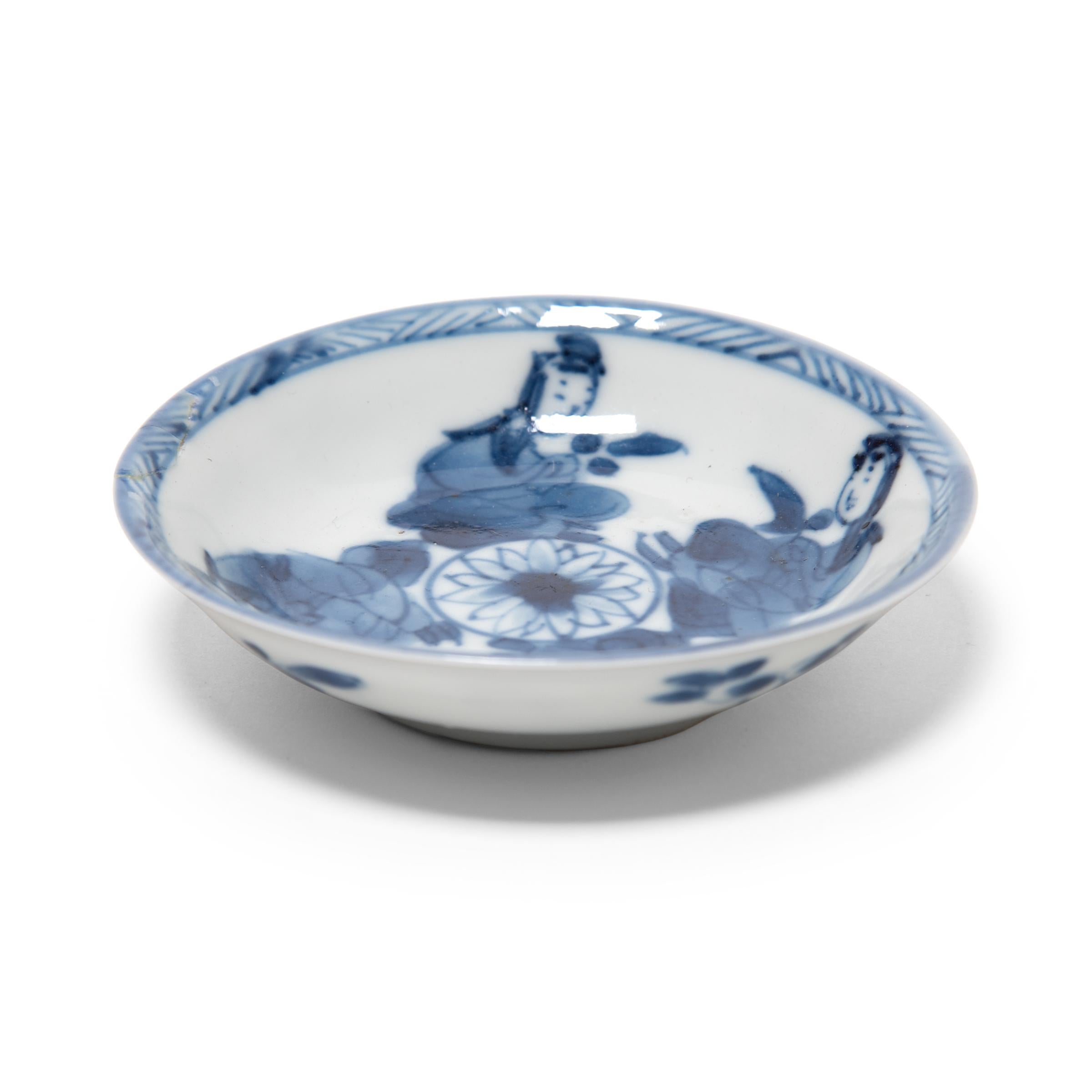 Diese Porzellanuntertasse aus dem 19. Jahrhundert ist ein schönes Beispiel für chinesische Blau-Weiß-Keramik. Die zierliche Schale ist fein gearbeitet mit dünnen Wänden und zarten Pinselstrichen, die vier um eine zentrale Lotusblüte sitzende Diener
