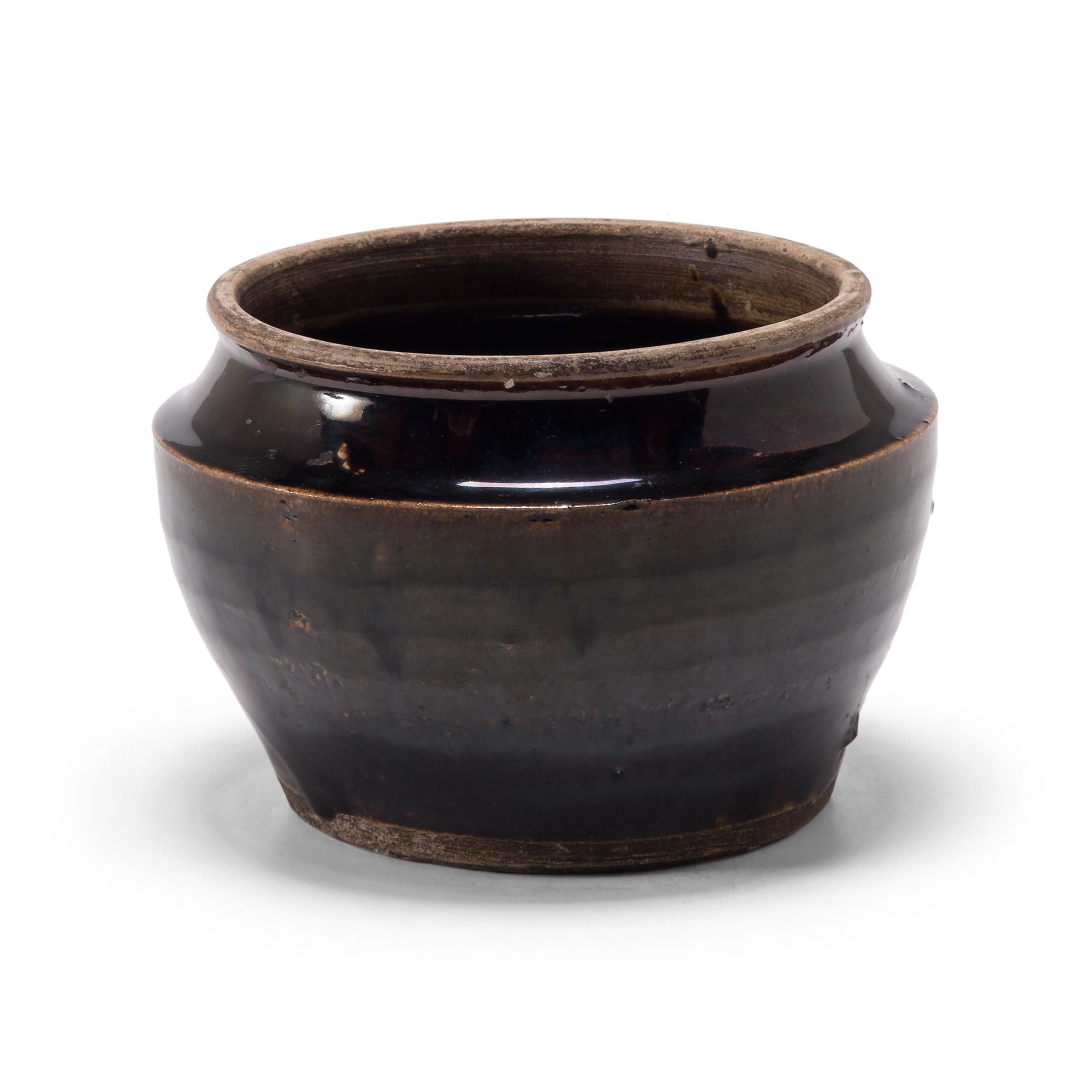 A dark glaze coats the angular body of this squat kitchen vessel, creating a subtle gradient as it spills over the tapered sides. The petite early 20th century dish was once used for storing food in a Qing dynasty kitchen, as evidenced by its