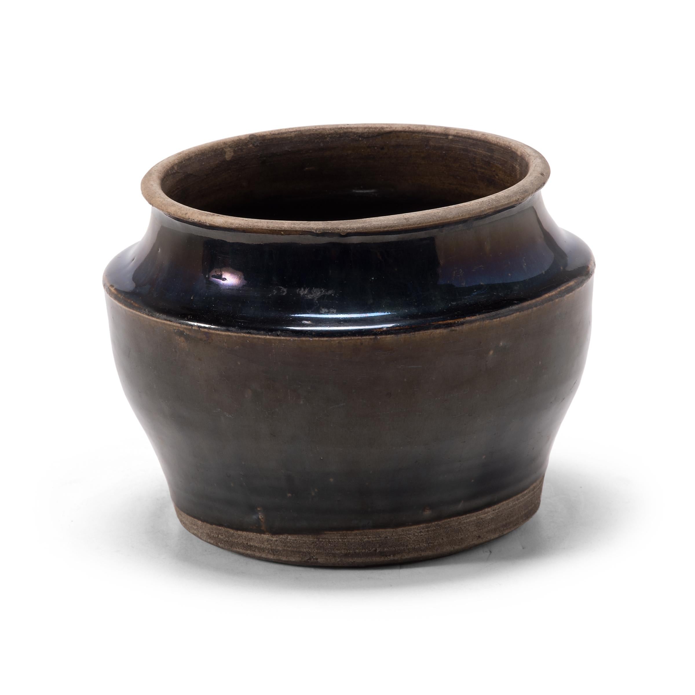 A dark glaze coats the body of this squat kitchen vessel, pooling at the angular shoulders. The petite early 20th century dish was once used for storing food in a Qing-dynasty kitchen, as evidenced by its interior glaze. Dotted with imperfections,
