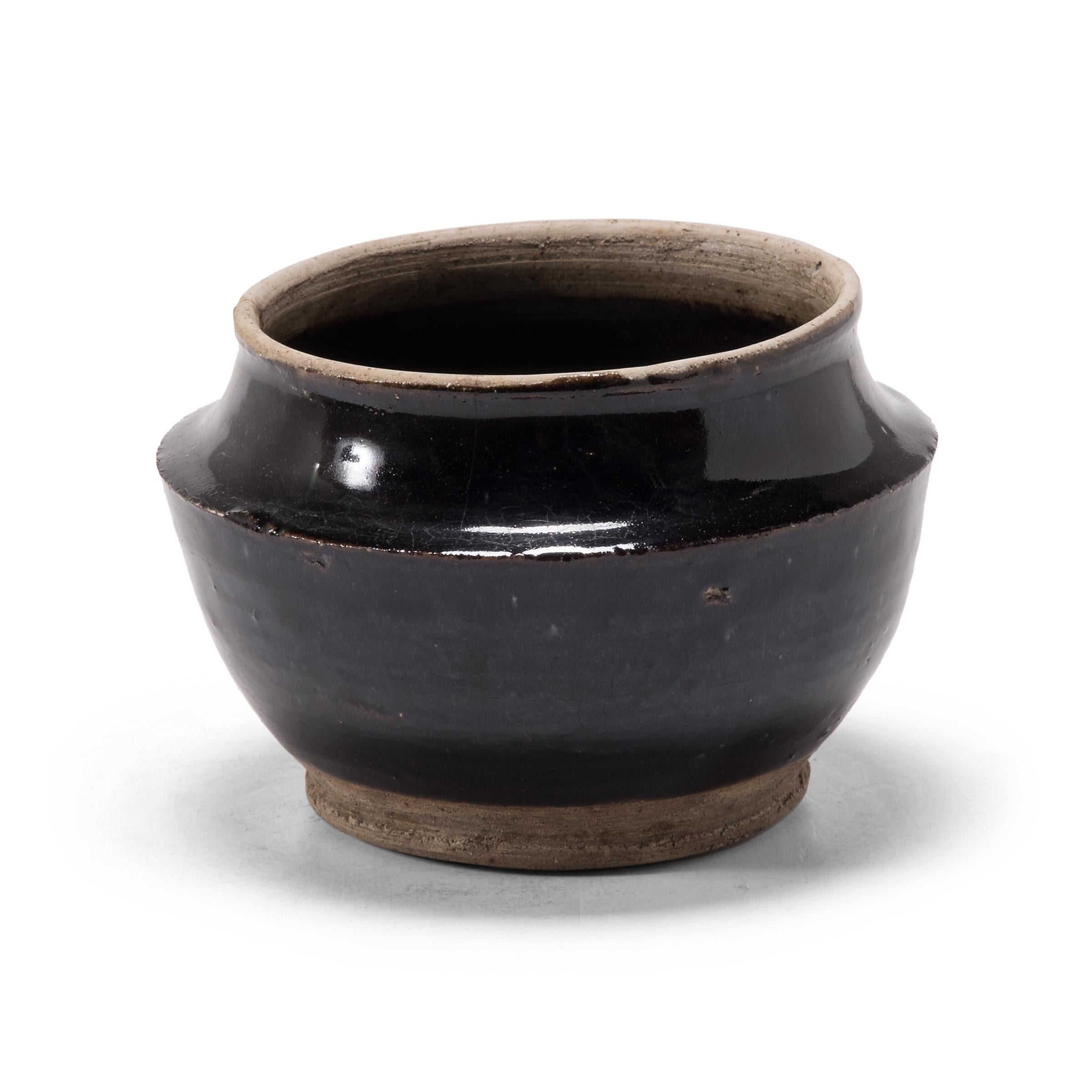 A dark glaze coats the body of this squat kitchen vessel, pooling at the angular shoulders. The petite early 20th century dish was once used for storing food in a Qing-dynasty kitchen, as evidenced by its interior glaze. Dotted with imperfections,