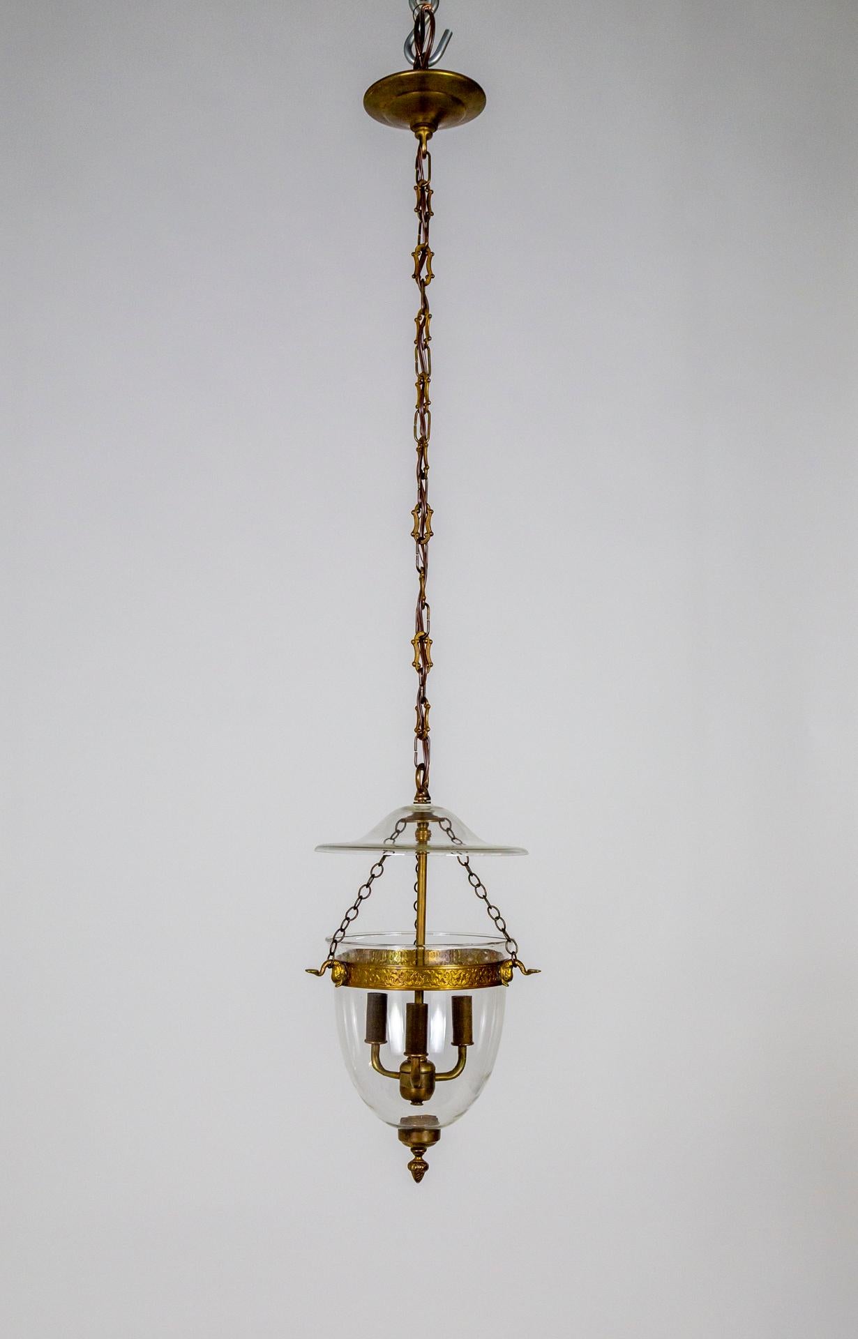 A traditional style bell jar light fixture with a glass smoke cap and brass parts toned in bronze and gold. The rim is pressed with an acanthus leaf pattern and held with cast dolphins to the chains. It hangs from a long, decorative chain and