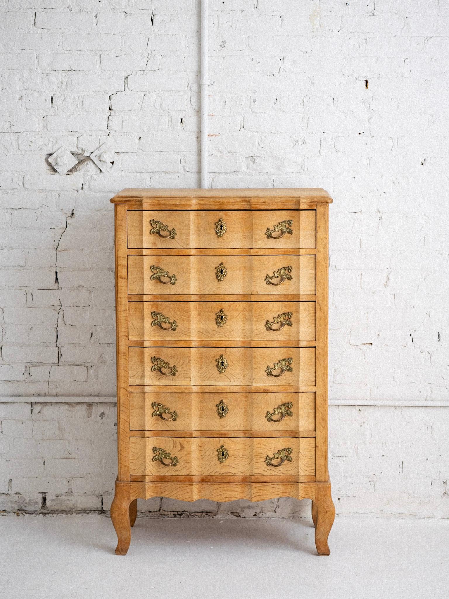 A petite mid century Danish tallboy. In the manner of Henning Kjaernulf. Blonde oak finish and brass hardware. Serpentine sculptured silhouette. Six locking drawers. Key included. Upper drawer includes a sliding accessory tray.