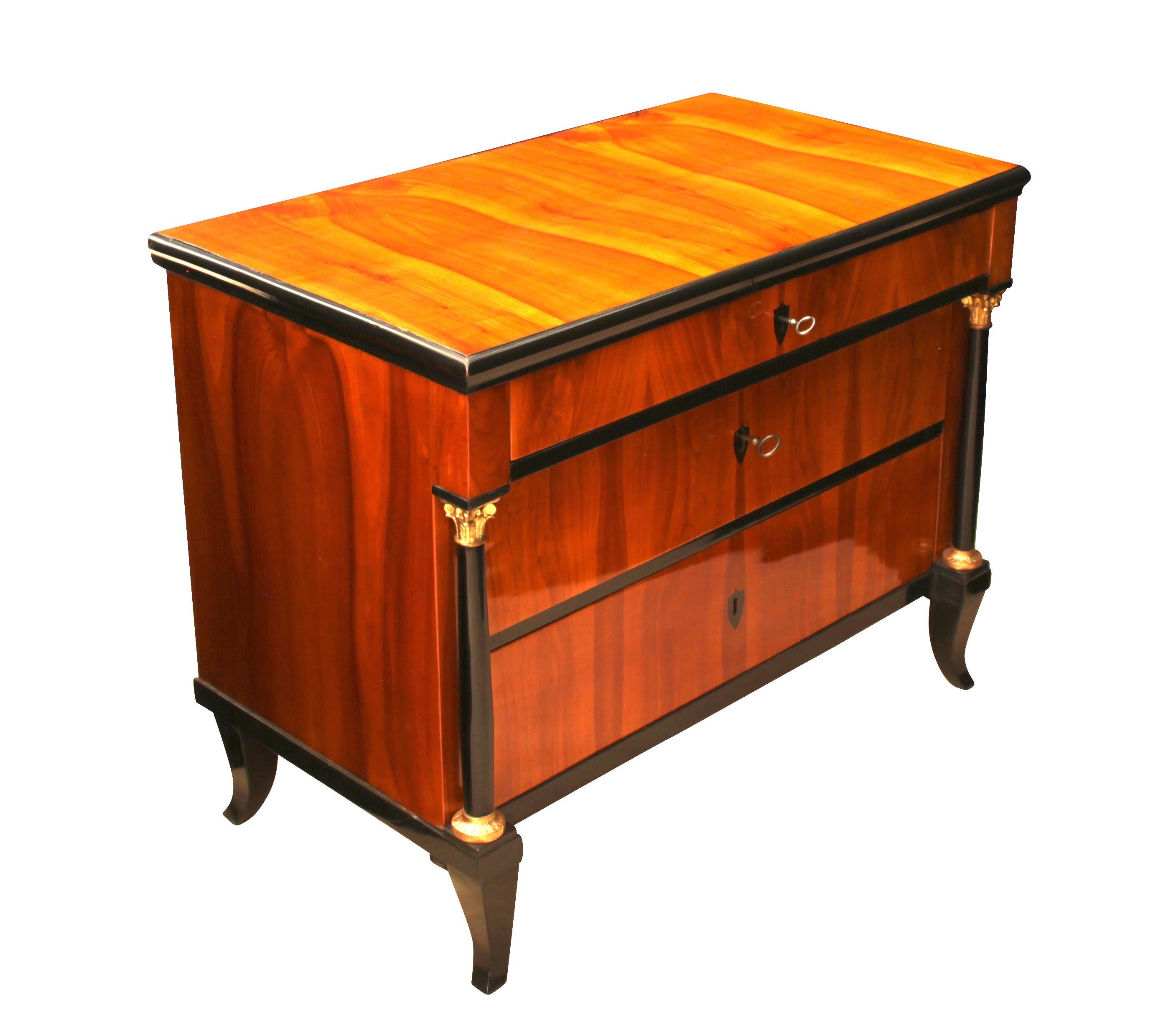 Very elegant, petite original Commode/Chest of Three Drawers from about 1815, marking the transition from Empire to early Biedermeier. 
It has two black bottle-shaped full-columns with gold-plated corinthian capitals and bases. The beautiful bright