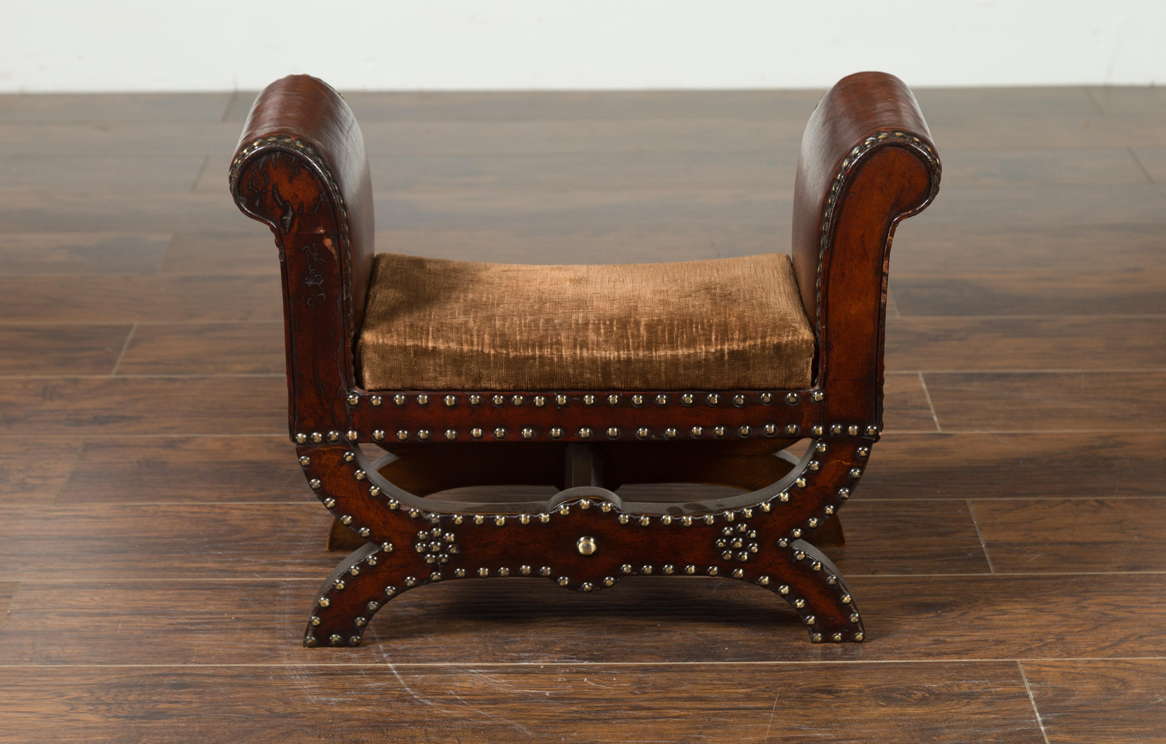 A petite English leather-bound stool from the early 20th century, with out-scrolling arms and nailheads. Created in England during the first quarter of the 20th century, this stool charms our eyes with its petite proportions and graceful silhouette.