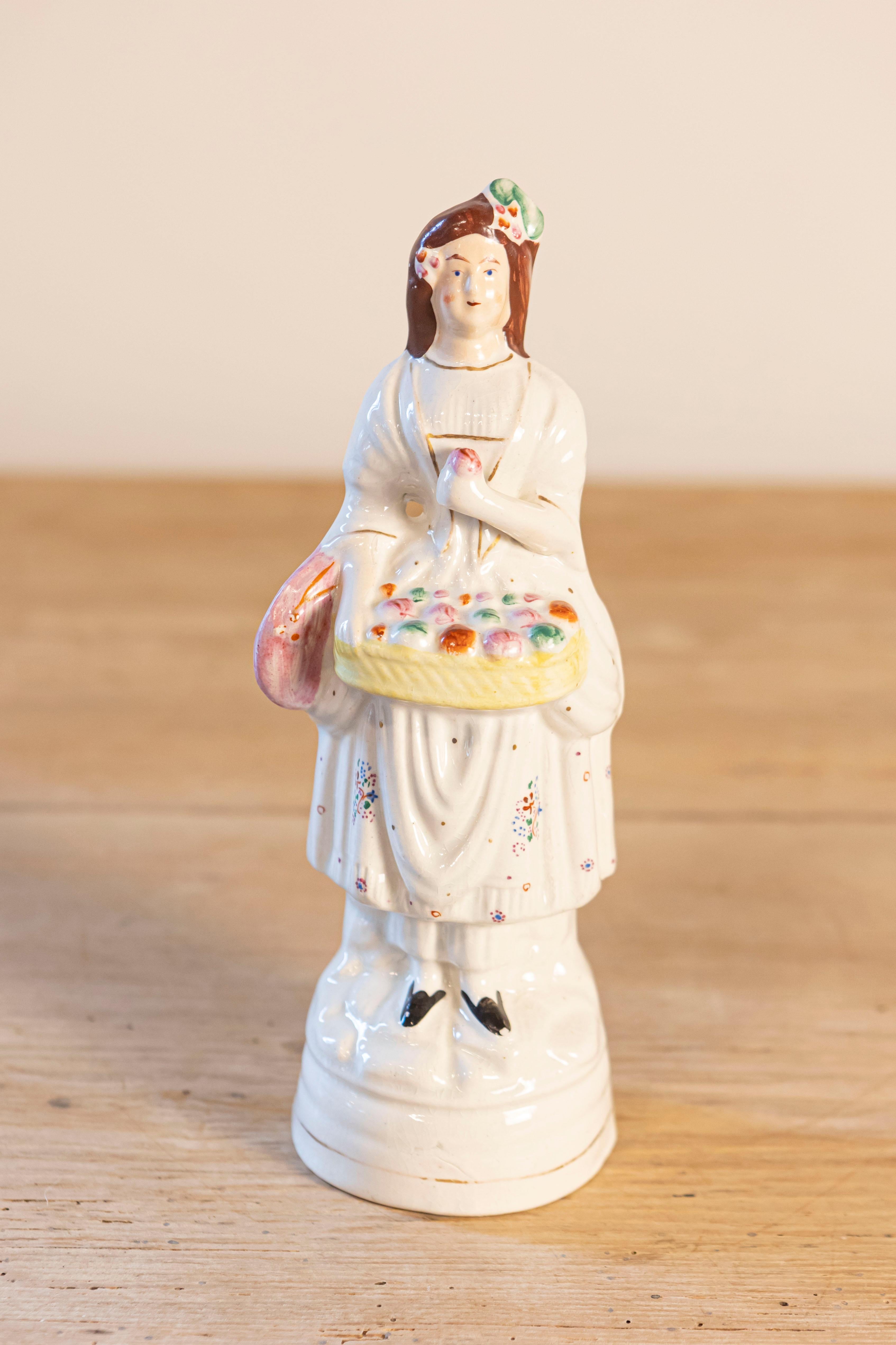 A small English porcelain decorative object depicting a lady carrying a floral basket. Created in England during the 20th century, this petite sculpture depicts a young lady, dressed in white with gilt trims, carrying a yellow wicker basket filled