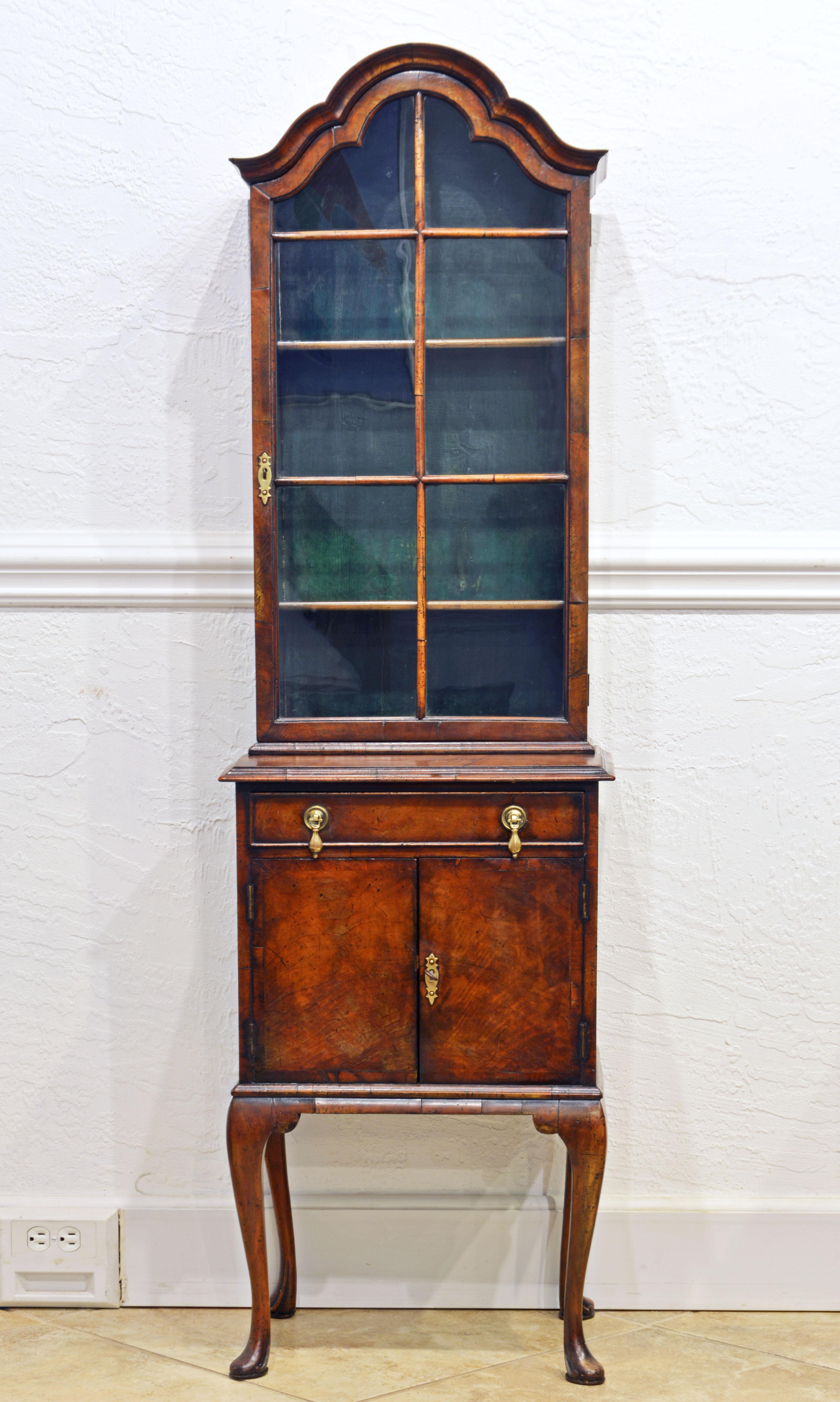 This adorable petite Queen Anne style burled walnut display or curio cabinet features a shaped domed top above two glazed doors with rounded mullions opening up to a painted and shelved interior. The lower part has a drawer above two doors opening