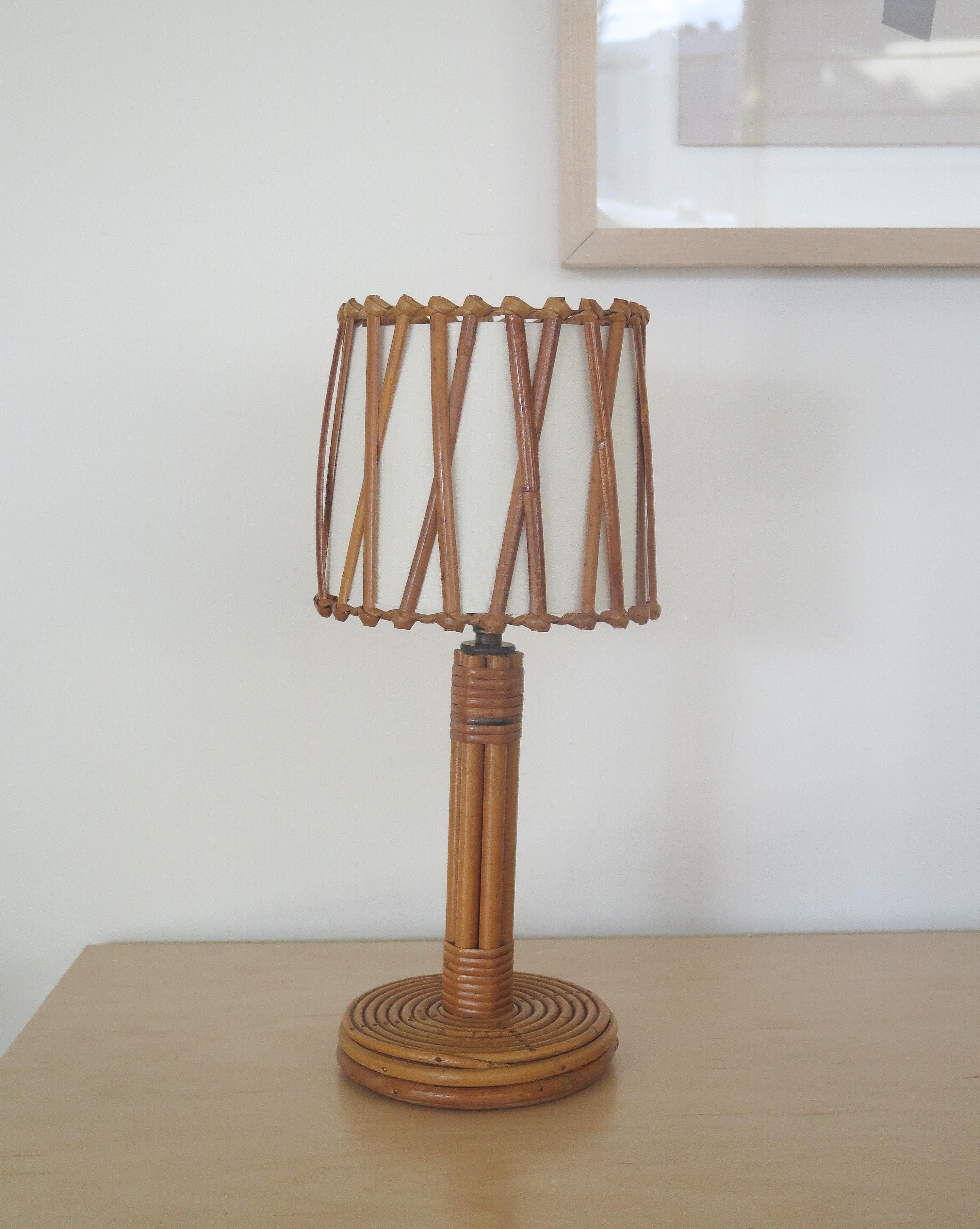 Petite French rattan table lamp in the style of Louis Sognot. Circular base and stem with rattan drum shade and criss-cross rattan detail and silk interior shade lining.



  