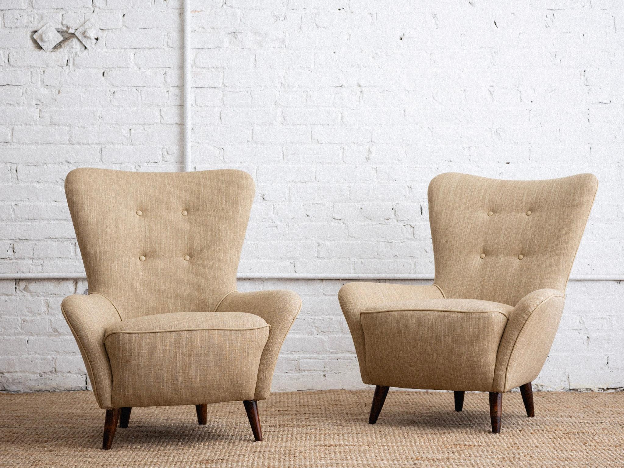 A pair of petite mid century Italian armchairs. Low profile wingback silhouette. Newly upholstered in a beige linen blend and matching leather trim. Sourced in Northern Italy.