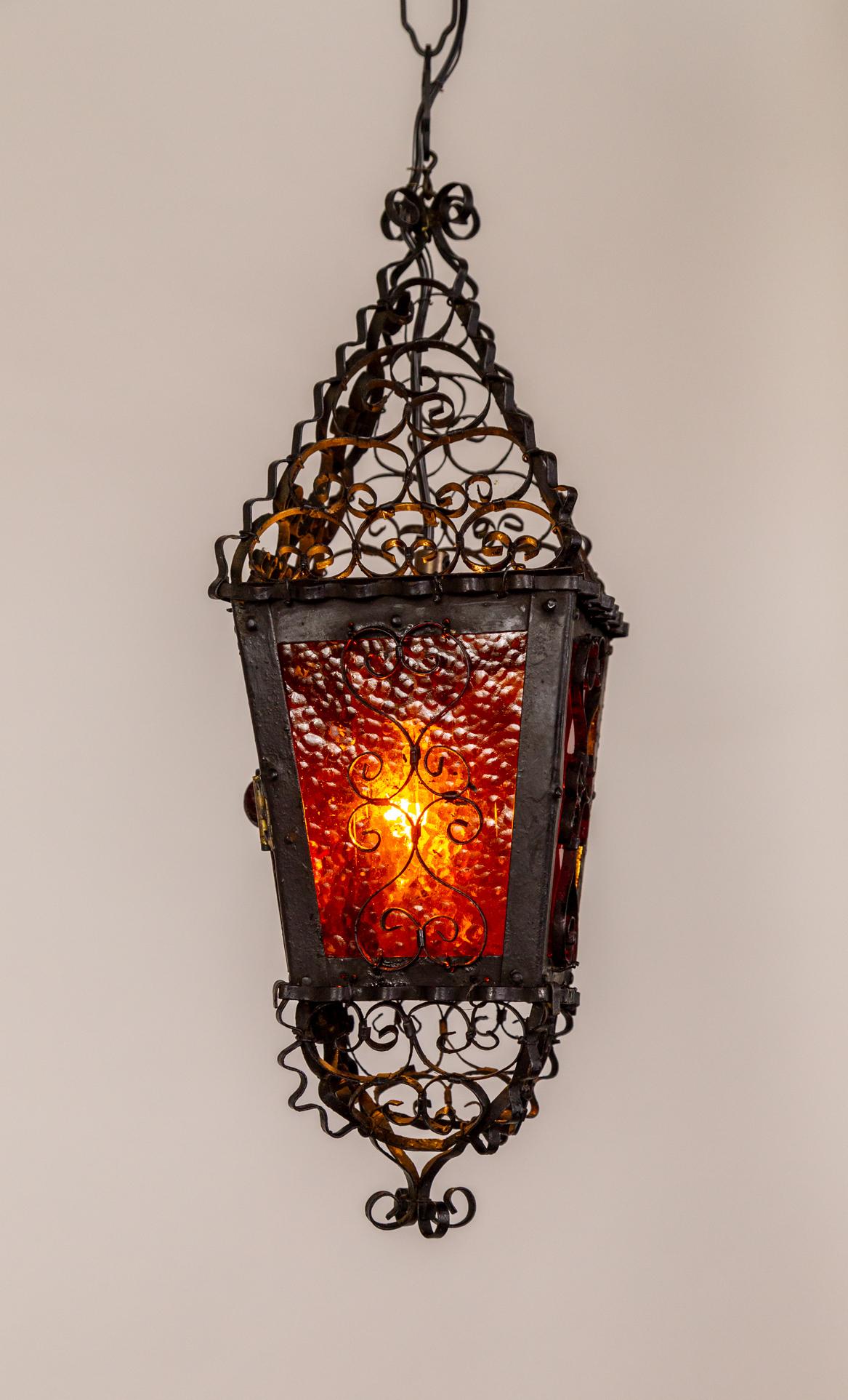 A small hanging lantern with deep orange and red glass panels, handmade in a tapered, four-sided frame with an abundance of delicately cured wrought iron ribbons clustered on the top and bottom.  It holds a single candelabra bulb. Newly rewired with