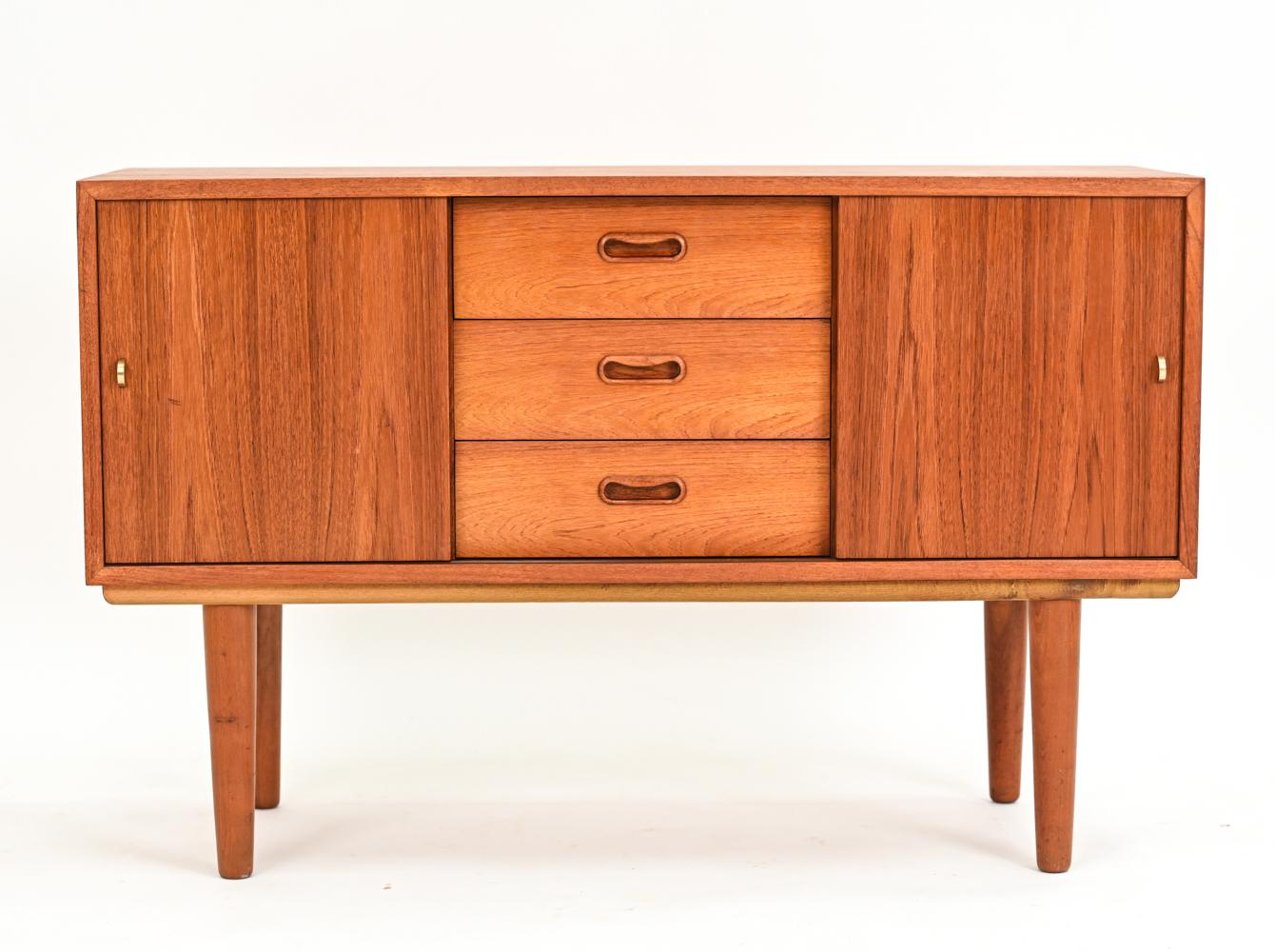 A diminutive apartment-scale sideboard or low chest in the manner of Kai Kristiansen. This charming Danish mid-century cabinet features three dovetailed drawers with shaped recessed pulls flanked by two sliding doors with brass half-moon handles.