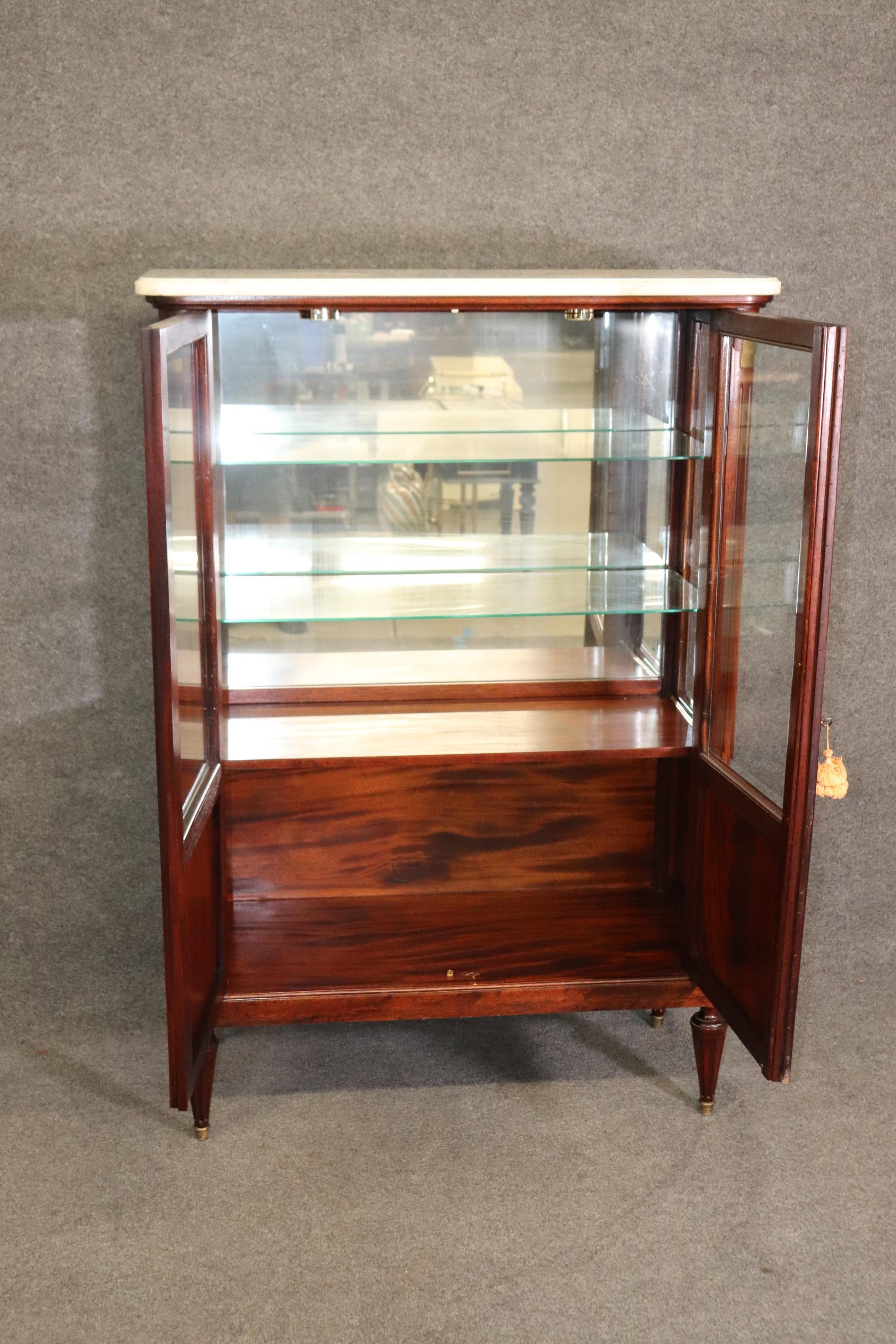 This is a great, smaller sized apartment style flame mahogany lighted vitrine or china cabinet. It has beautiful flame mahogany and a nice white marble top and brass accents. The cabinet dates to the 1940s and measures 60 tall x 41.25 wide x 18