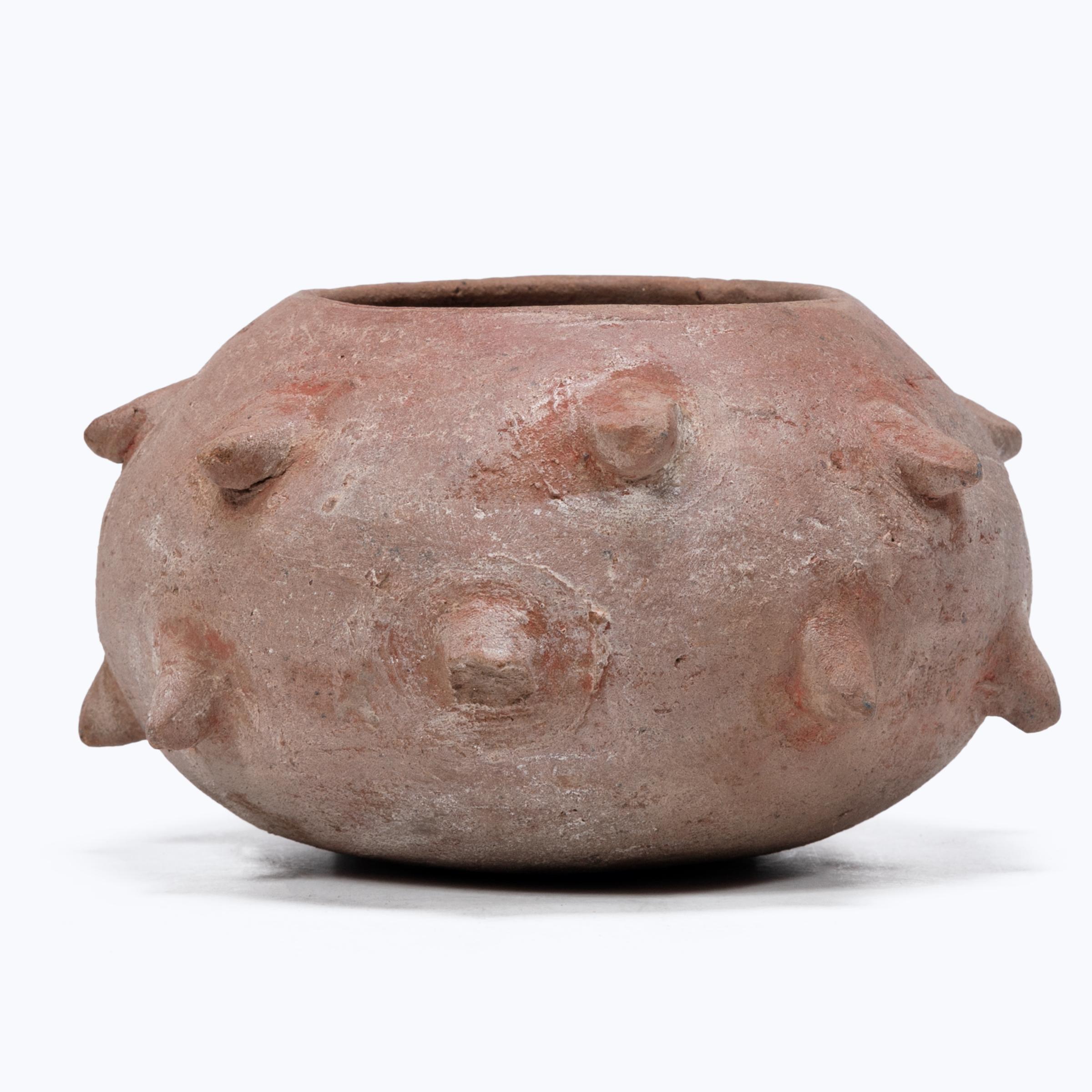 Once cloaked in a vibrant red clay slip, this beautifully worn petite vessel is actually a Maya censer. Molded spikes affixed to the exterior resemble those found on the Ceiba tree, the world tree of the Maya representing the universe and serving as