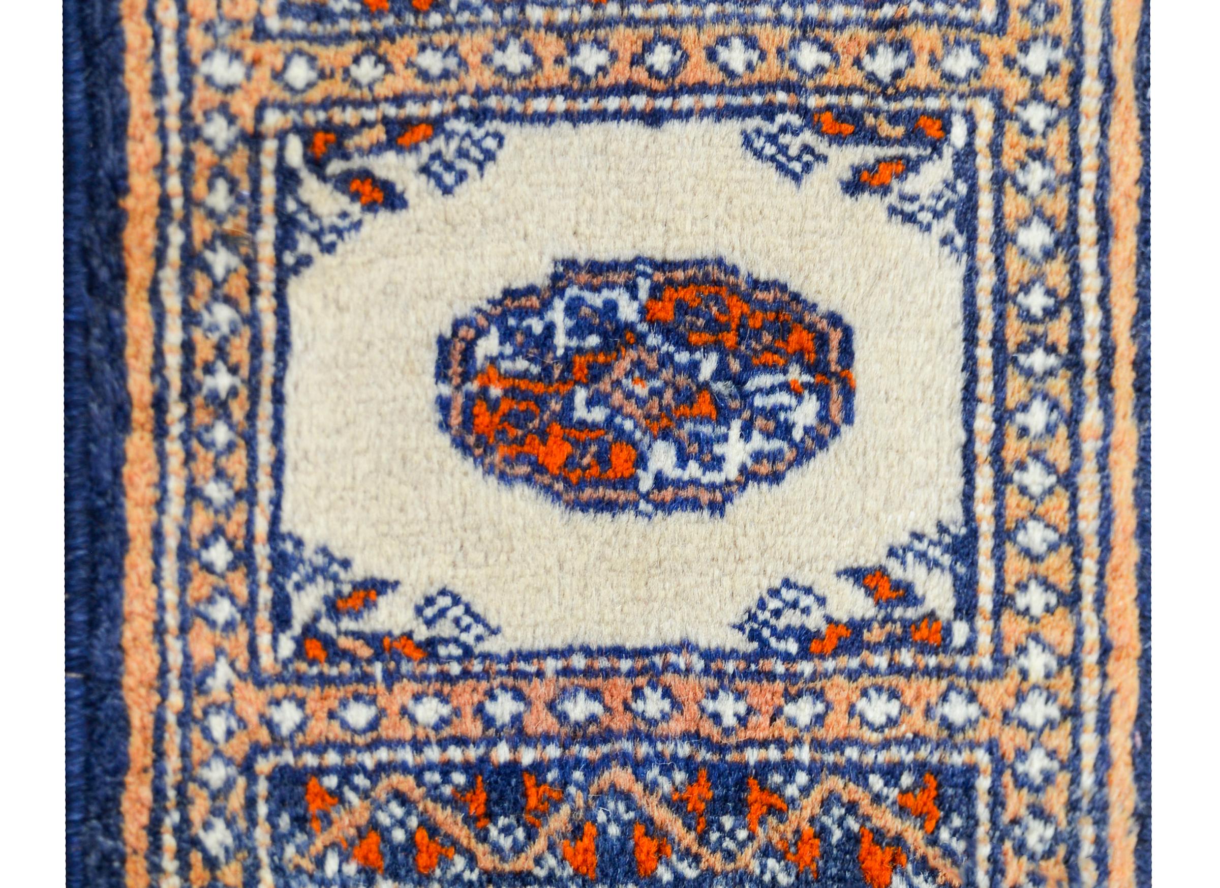 A petite vintage Pakistani Bakhara rug with a simple central medallion woven in orange and white, set against a cream background, and surrounded by border of geometric patterns woven in similar colors as the field.