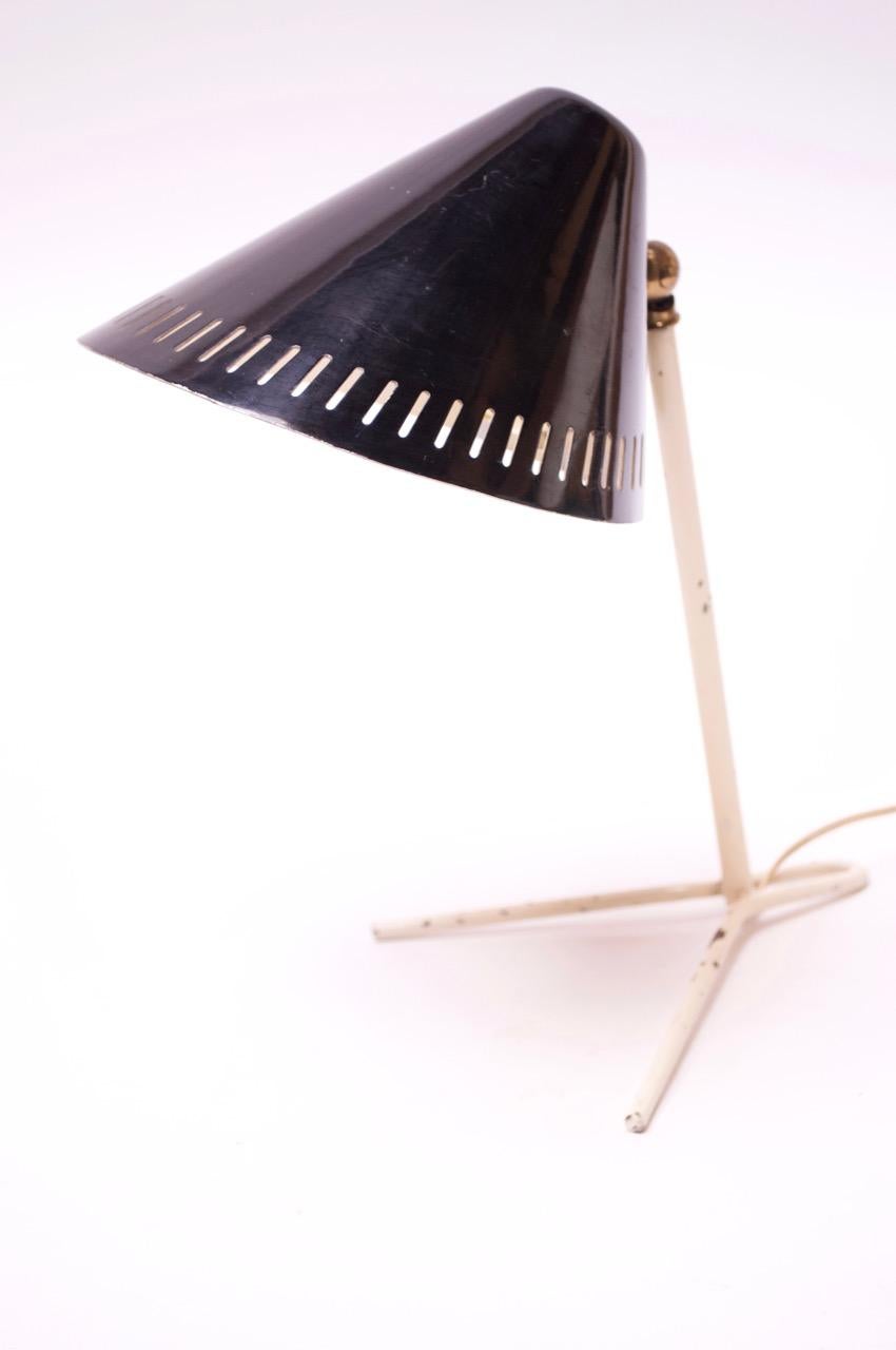 Charming, petite Italian table lamp (circa 1950s) composed of an incised black metal shade and off-white splayed-leg base. A brass ball joint connects the shade to the base and additionally allows the shade to be adjusted upward and downward. Lamp