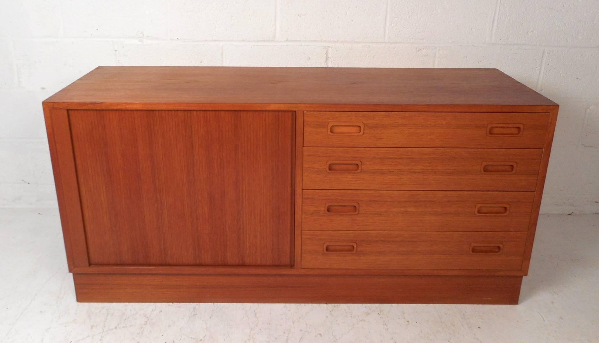 Questa bellissima credenza moderna vintage è caratterizzata da un'anta a tamburo che nasconde un ampio vano con due ripiani e quattro grandi cassetti. Design elegante con interni di colore biondo ed eleganti venature del legno di teak. Artigianato