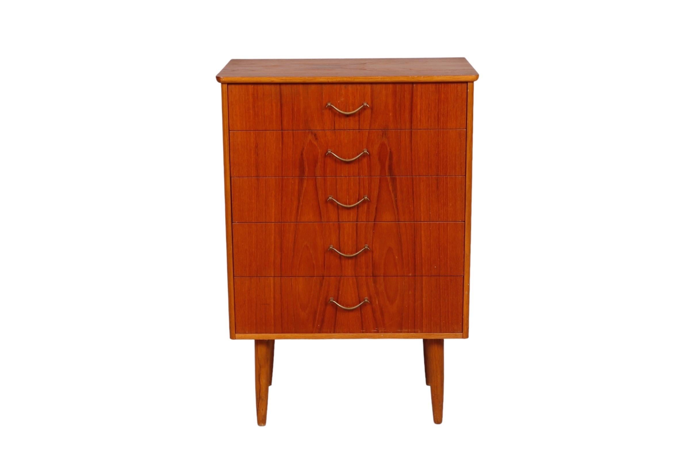 A petite mid-century dresser made of teak. Five drawers open with brass bail handles. Stands on round tapered legs.
