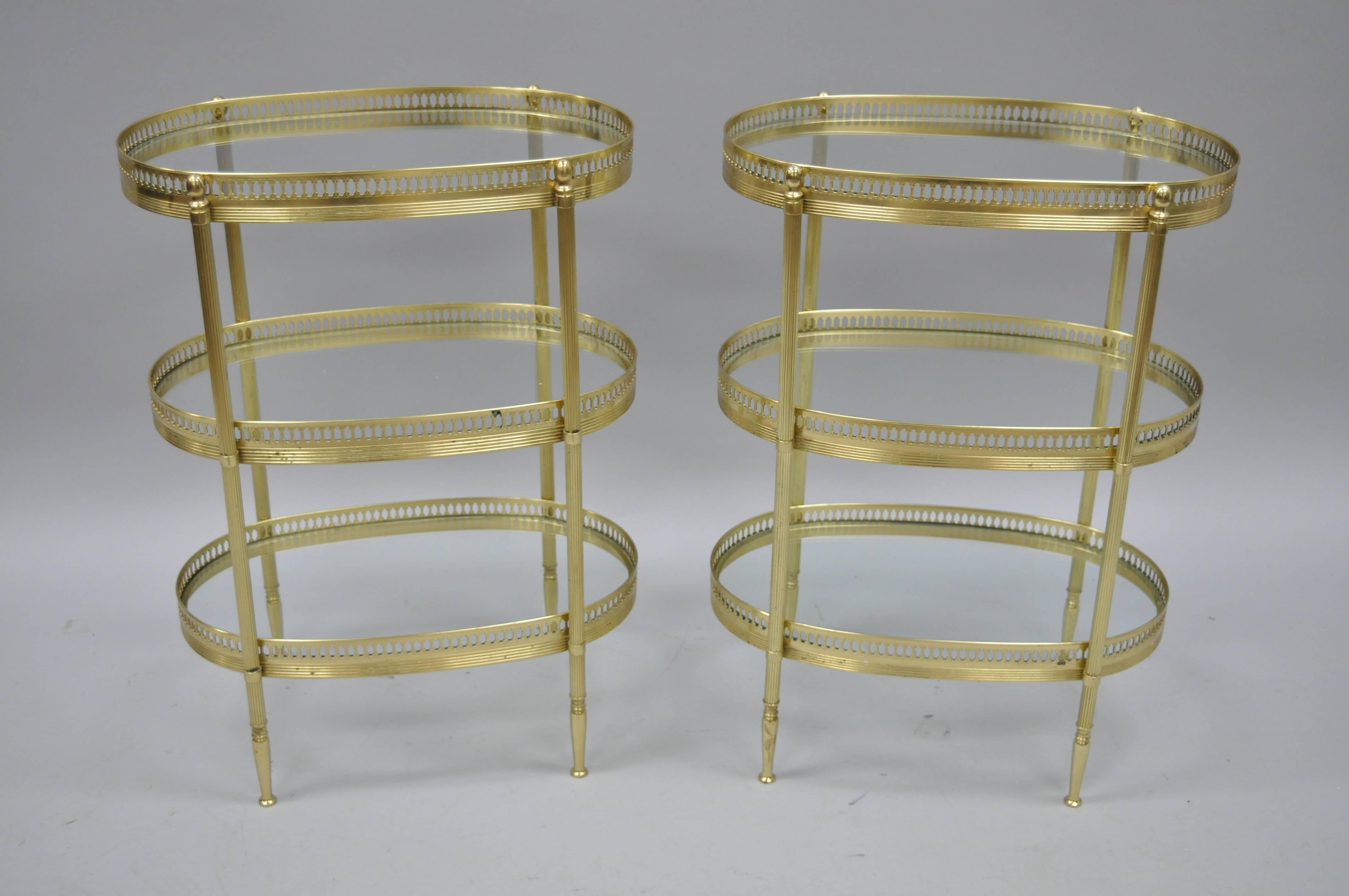 A vintage pair of petite brass oval end tables with inset glass shelves, circa mid-20th century. These Maison Jansen inspired French tables feature three tiers of clear glass shelves decorated with oval pierced galleries. Tables are raised on four