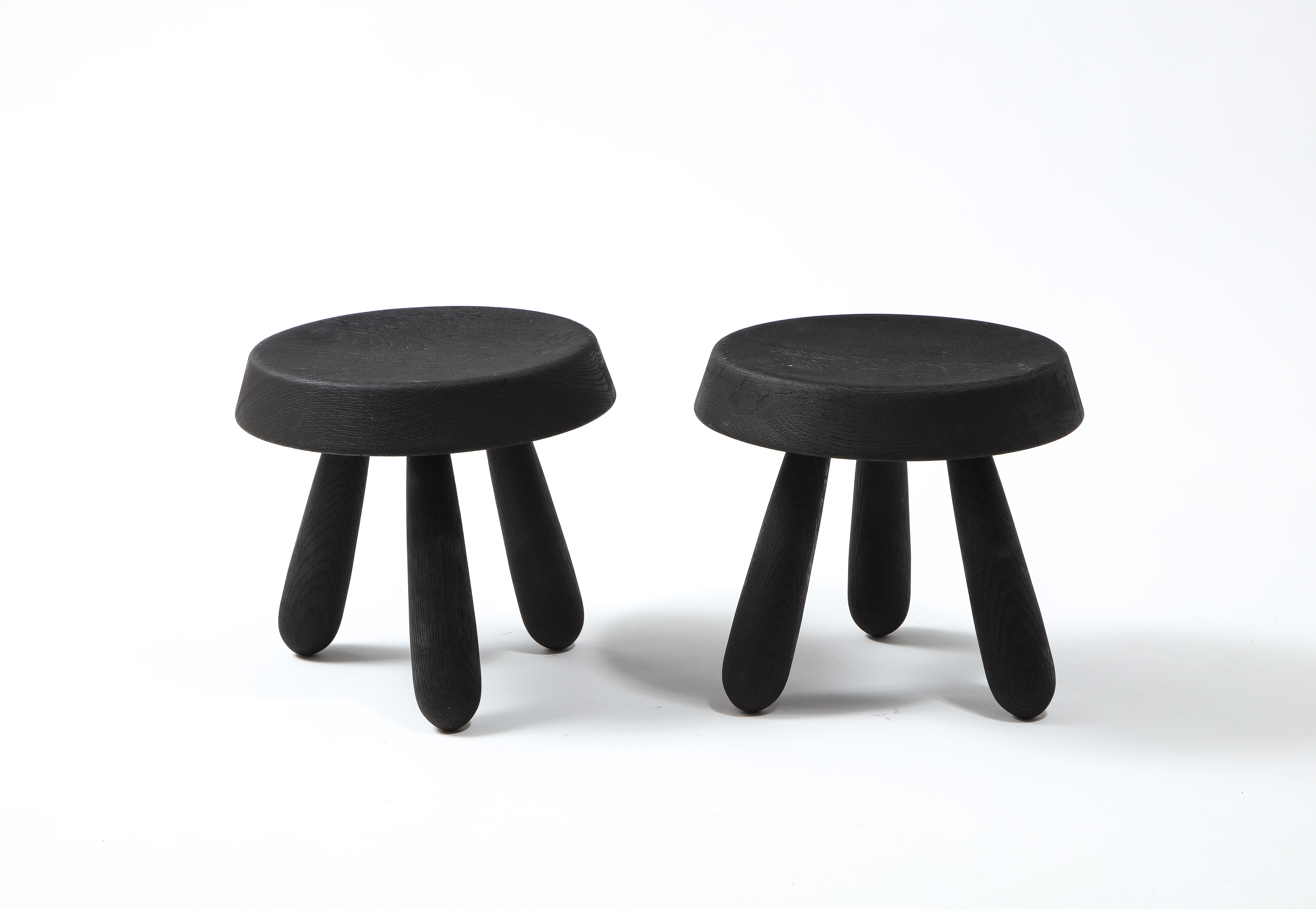 Pair of beautifully crafted petite tripod stools by French designer Douglas Mont. A thick beveled seat sits on three spindle shaped feet. Burnt Oak finish with visible wood grain. Appearance, cracks and grain may slightly vary as each stool is