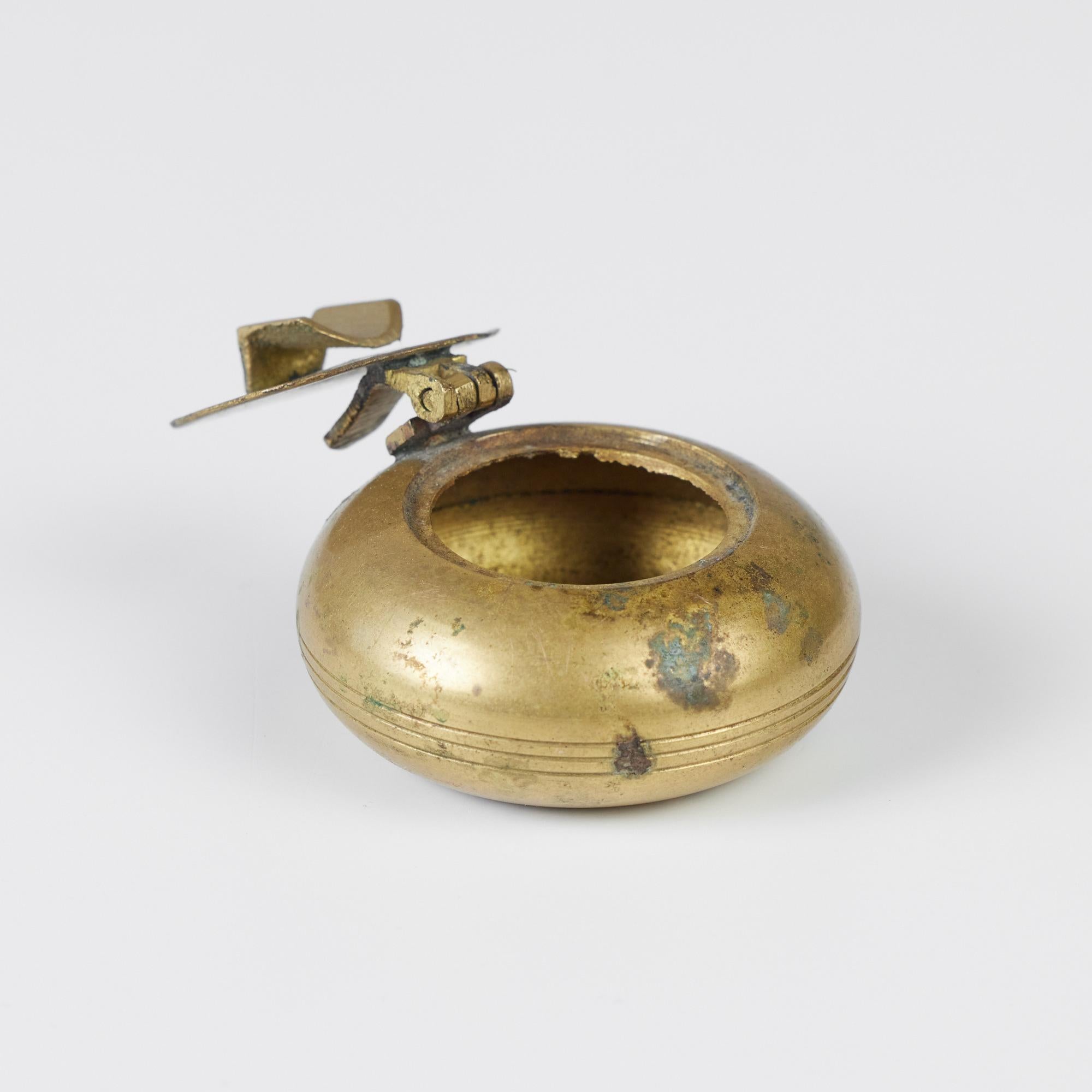 Petite brass ashtray with flip top lid. The ashtray features a patinated brass circular form with hints of verdigris and flip top opening. The circumference of the ashtray has an incised detail of three lines.

Dimensions: 2