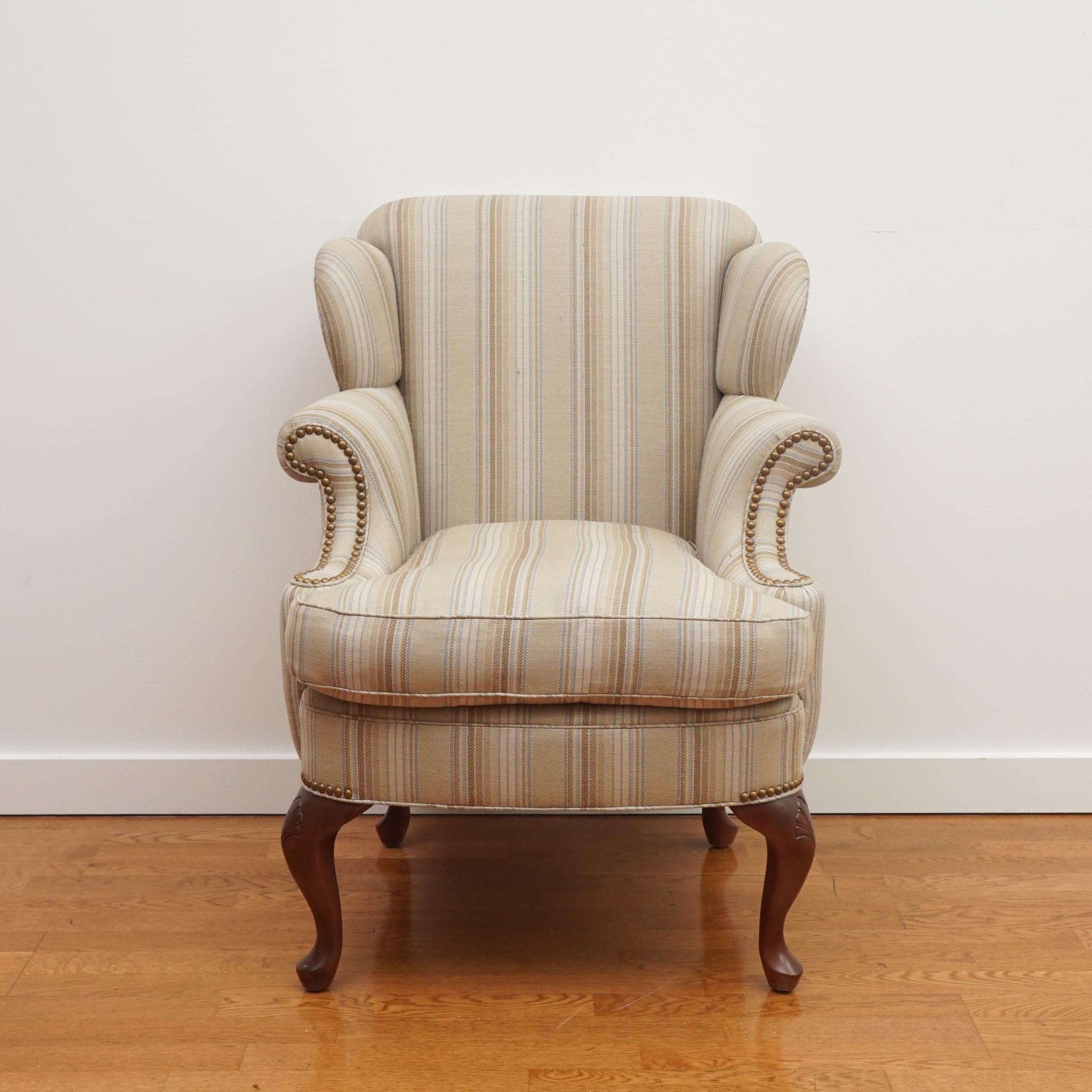 The petite Queen Anne-style wing chair, shown here, is desirable for both its scale and graceful style. Upholstered in a traditional multi-stripe fabric, the chair features carved cabriole legs in walnut finish and brass nail head detailing. The