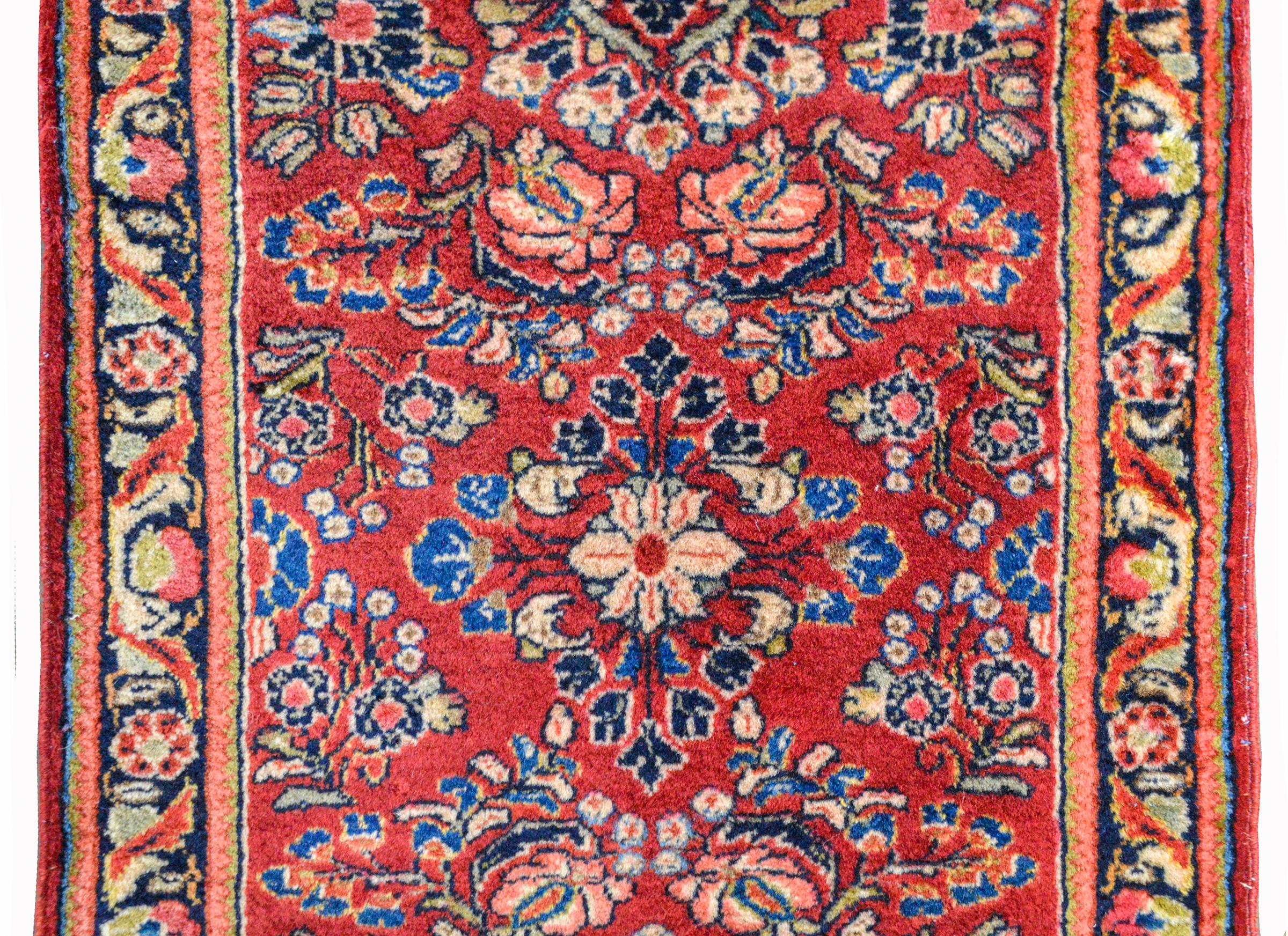 A patite early 20th century Persian Sarouk rug with a mirrored floral pattern woven in pink, indigo, cream, and green, against a dark cranberry background. The border is simple with a floral patterned central stripe flanked by simple geometric