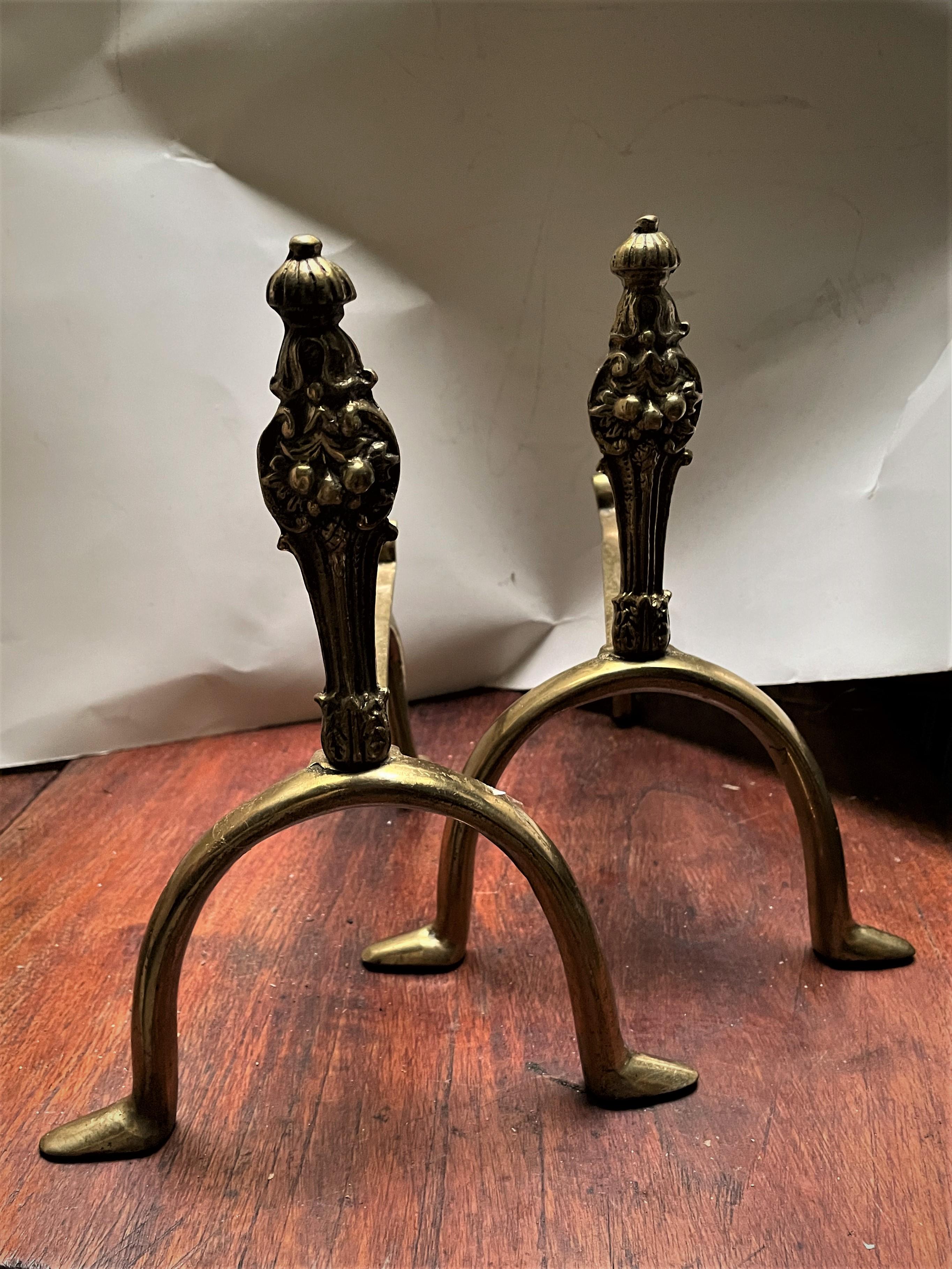 This is an exceptionally sweet pair of very small cast brass andirons. The plinth consists of acanthus leaves which raise to a decorative fruit grouping topped with a bell flower. The semi circle legs have very human looking feet facing sideways and