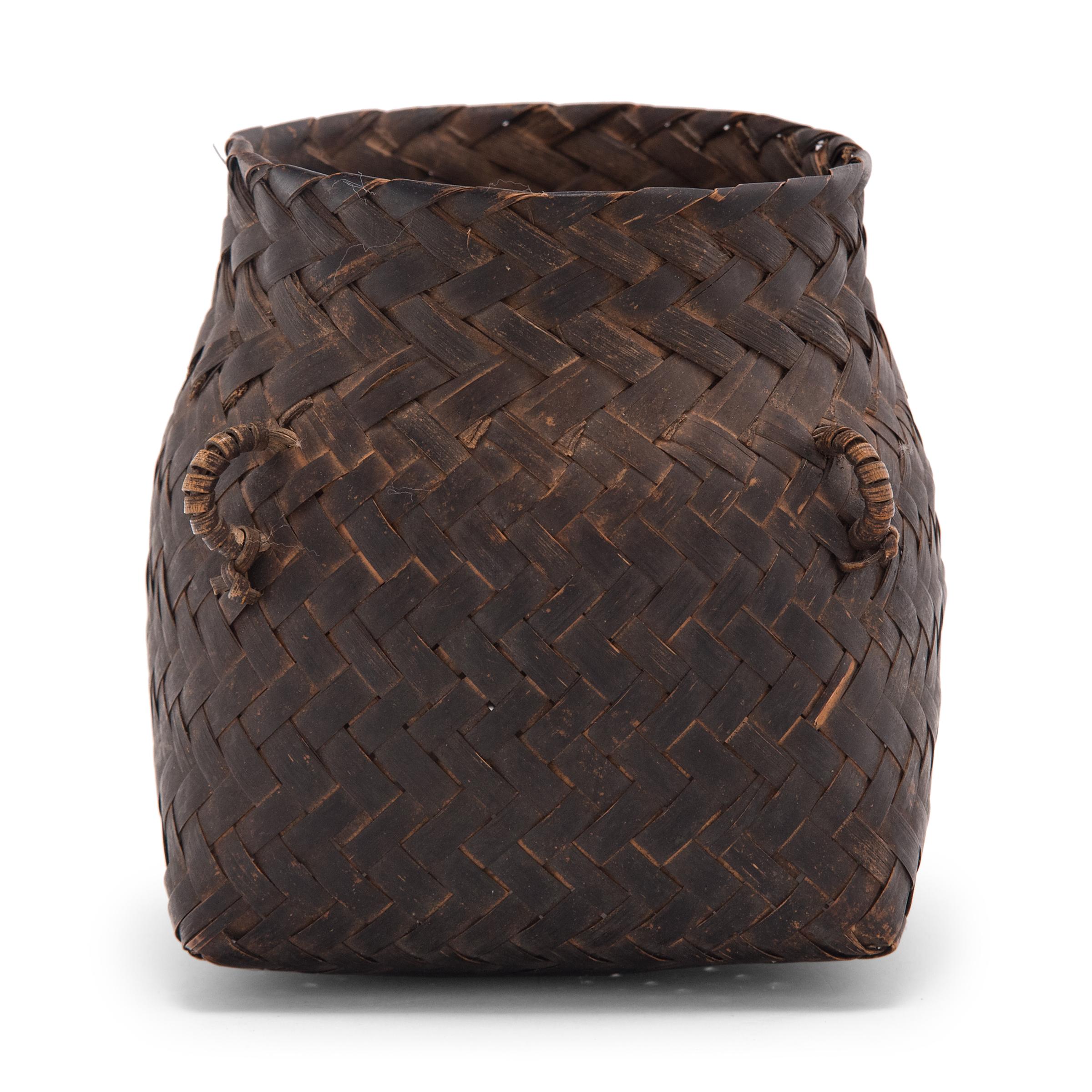 This petite woven basket dates to the early 20th century and is crafted of thin bamboo strips that have been carefully hand-woven in a twill pattern. The basket has a tapered form, narrowing from a square base to a round opening, and is darkened to